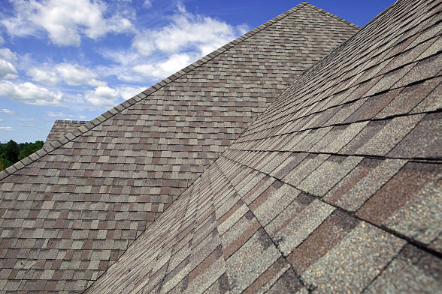 Close-up of a shingle roof under a blue sky with clouds, showing textured asphalt shingles.
