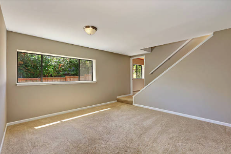 Empty room with beige carpet, beige walls, a window showing outdoor greenery, and a staircase. Roofing and painting in Bucks County