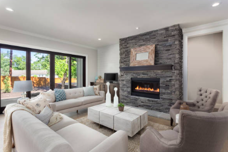 Modern living room with a fireplace, gray stone wall, beige couches, armchairs, and a wooden coffee table, featuring a large window.