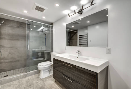 Modern bathroom with glass shower, white toilet, large mirror, dark wood vanity, and bright lighting.