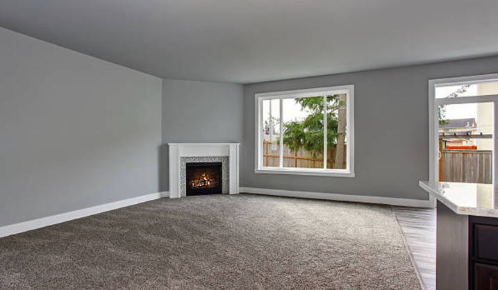 Modern living room with gray walls, a white fireplace, large window, beige carpet, and hardwood floor near kitchen counter. Roofing and painting in Bucks County