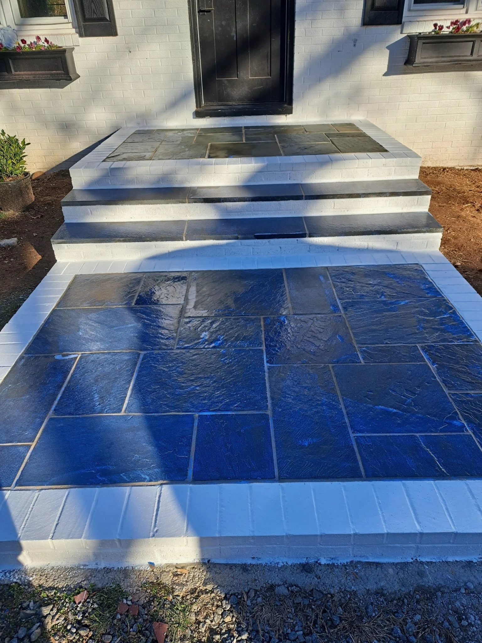 Front steps and porch of a house with black door and white brick exterior, with flower boxes on windows, blue-tiled landing, and white steps leading up to the door.