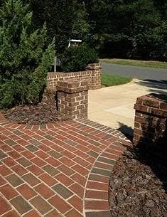 Brick pathway with a curved edge leading to a sidewalk and driveway, bordered by bushes and greenery.