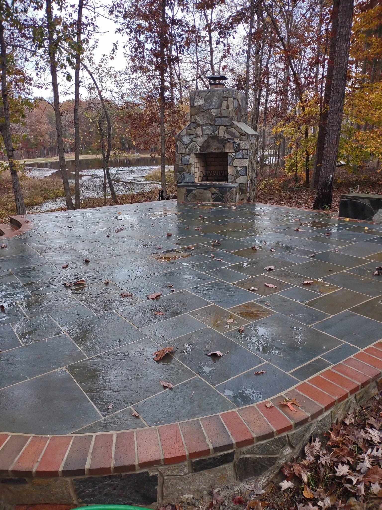 An outdoor stone patio with a fireplace in a wooded area, wet from recent rain, with fallen leaves scattered on the ground and the wet surface.