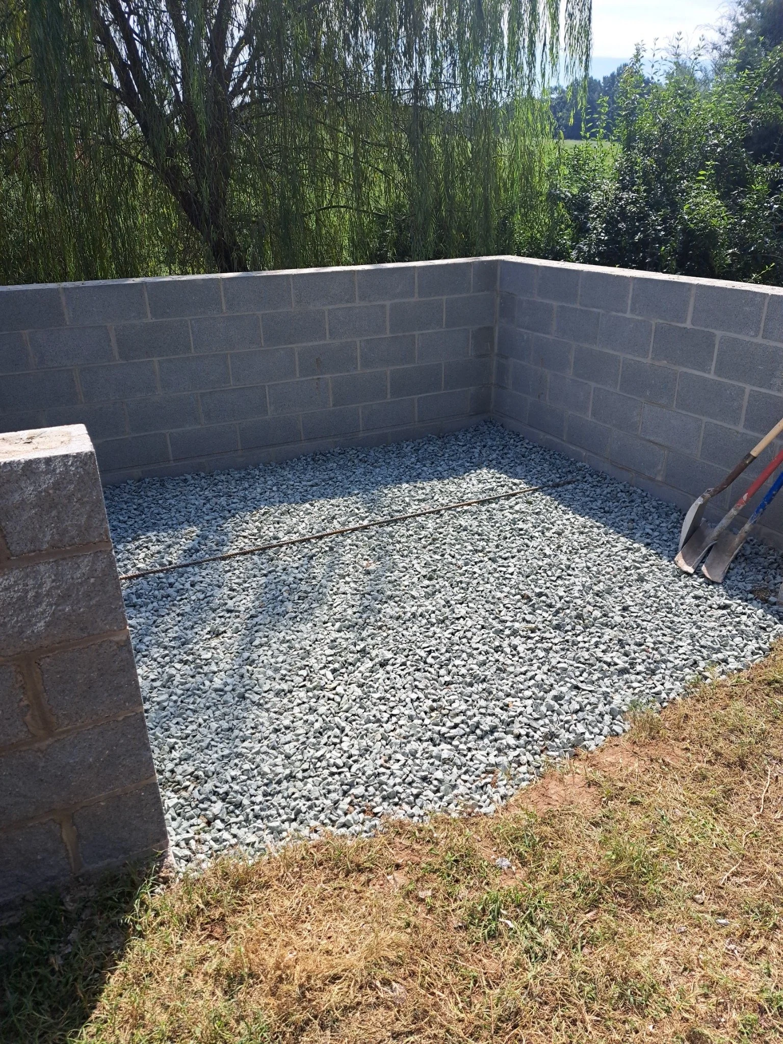 A small outdoor area with gravel ground, enclosed by gray cinder block walls, and shovels resting against the right wall. The surrounding area has grass and tall trees in the background.