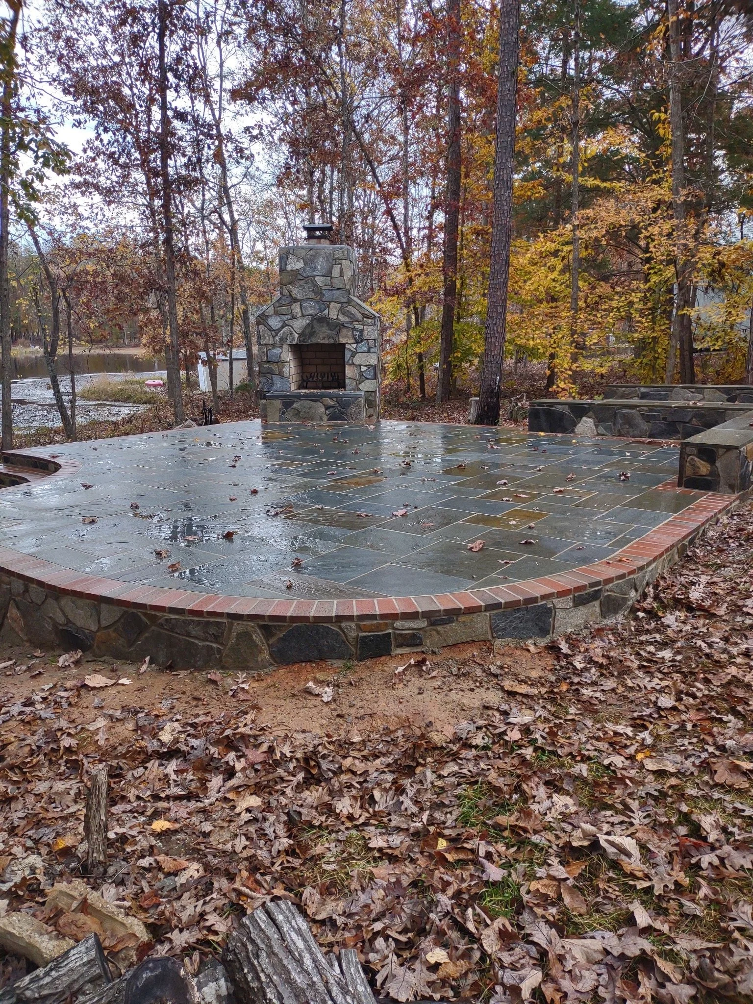 A stone and brick outdoor fireplace on a paved patio surrounded by fallen autumn leaves, with trees and a lake in the background.