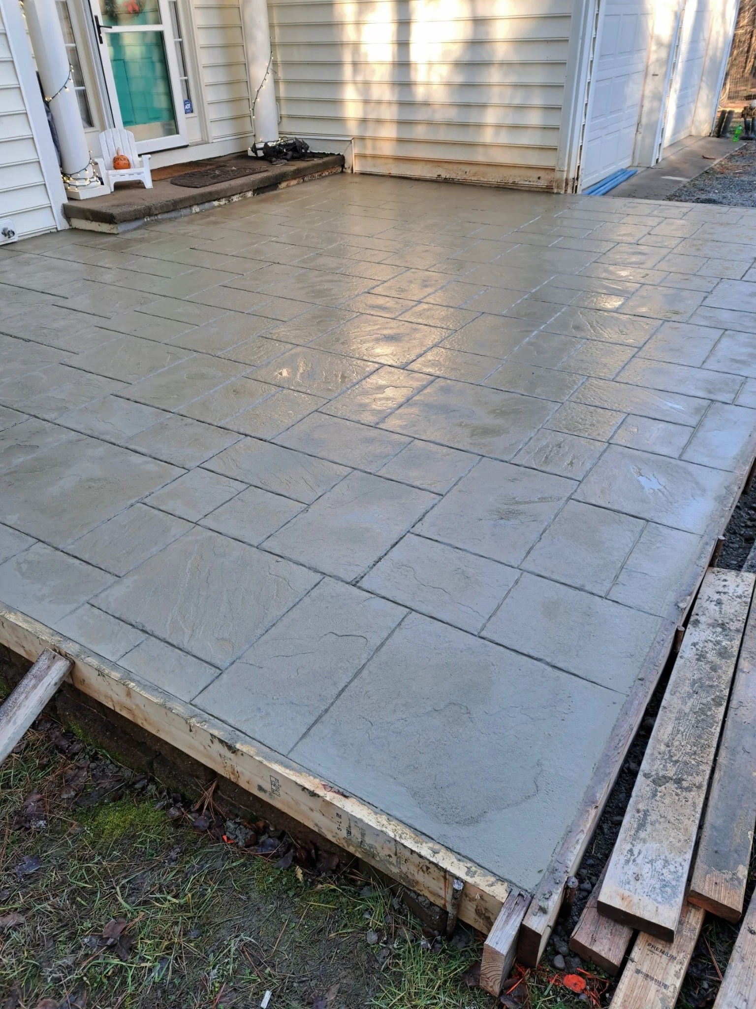 Recently poured concrete patio with decorative stamped finish, adjacent to house with vinyl siding, next to garage, with building supplies and wooden framework visible.