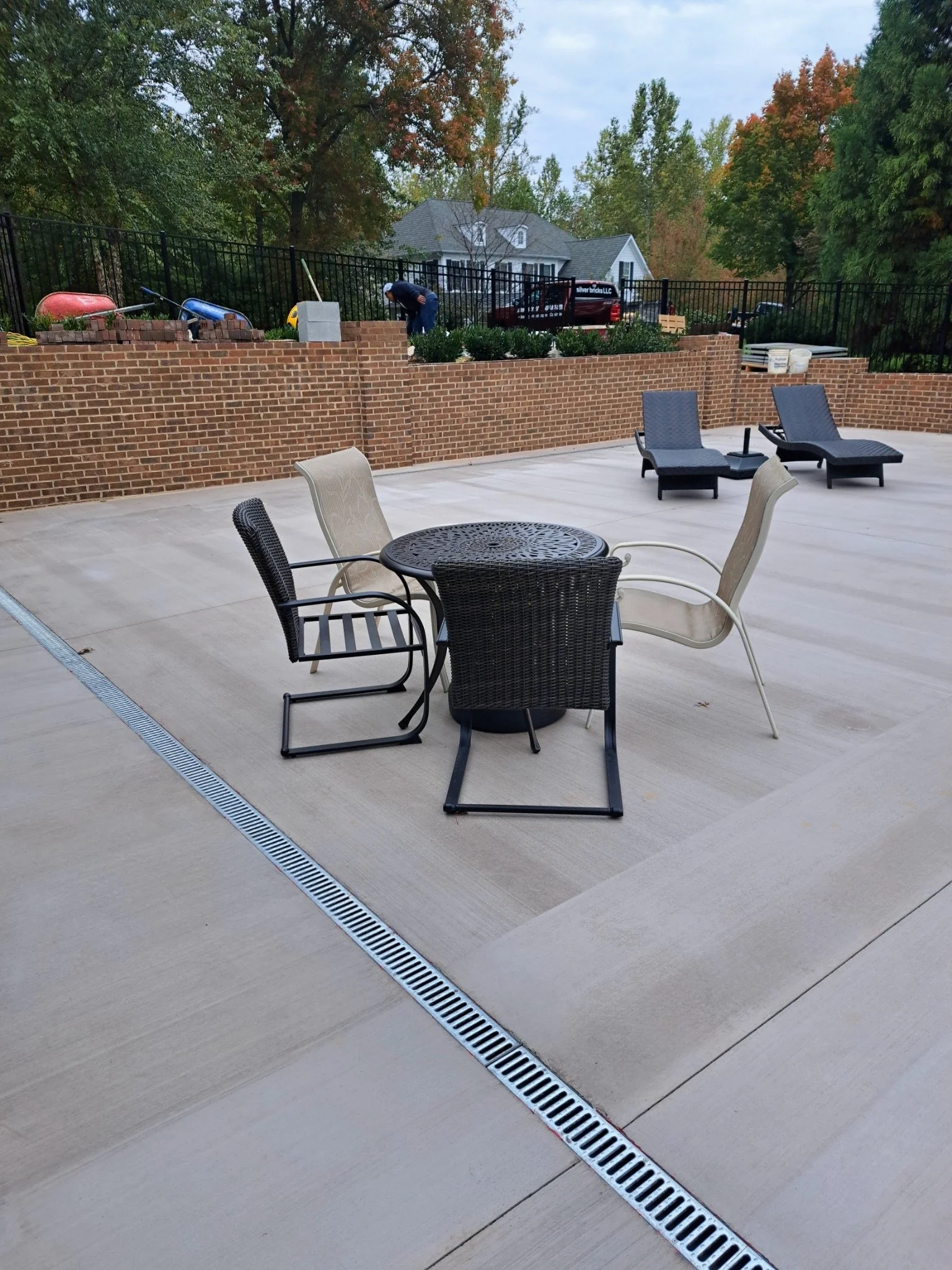 An outdoor patio with patio furniture, including a round table with four chairs and two lounge chairs, with a brick wall, trees, and a house in the background.