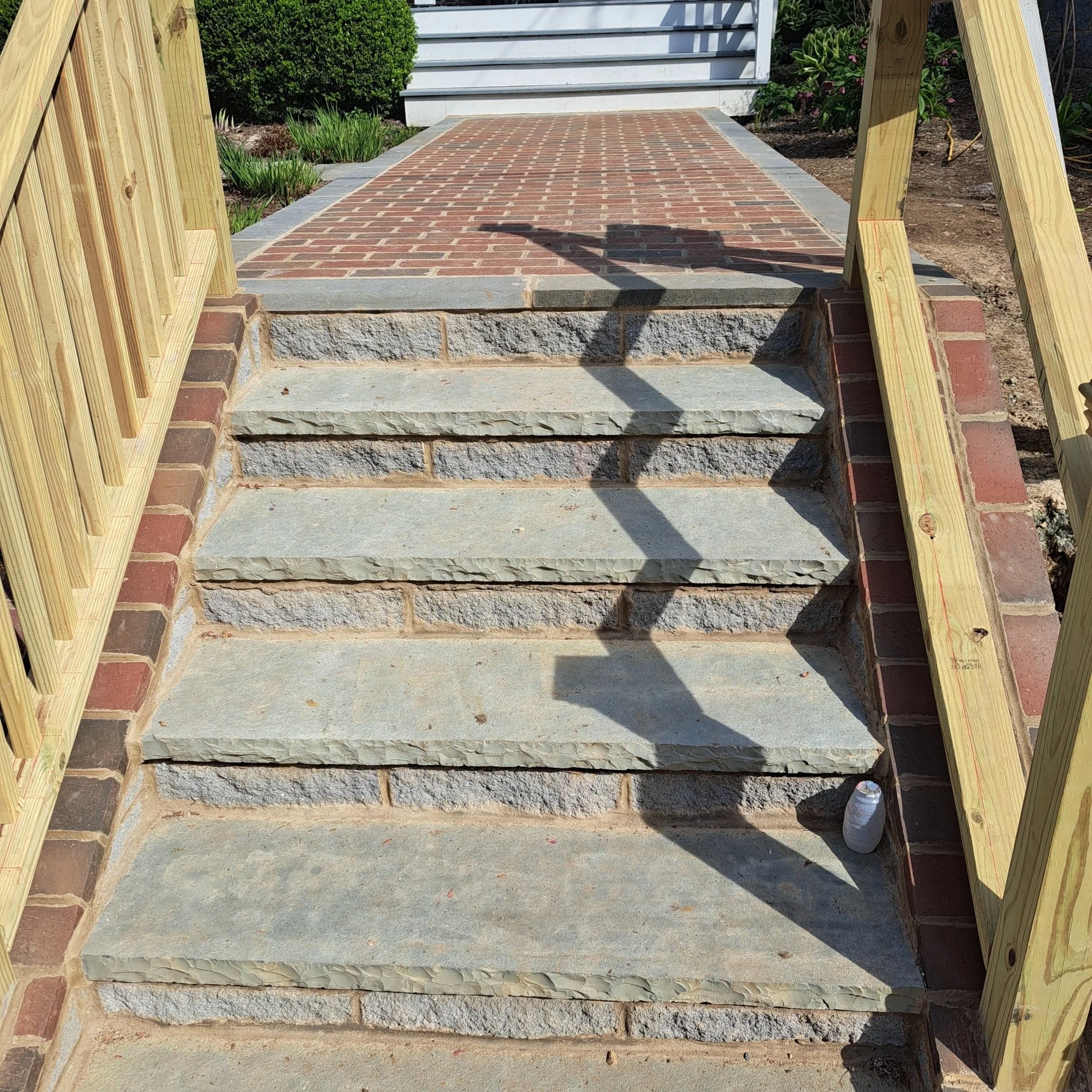 Concrete and brick steps leading up to a brick and stone porch, with a wooden railing on both sides and a shadow of a streetlamp cast on the steps