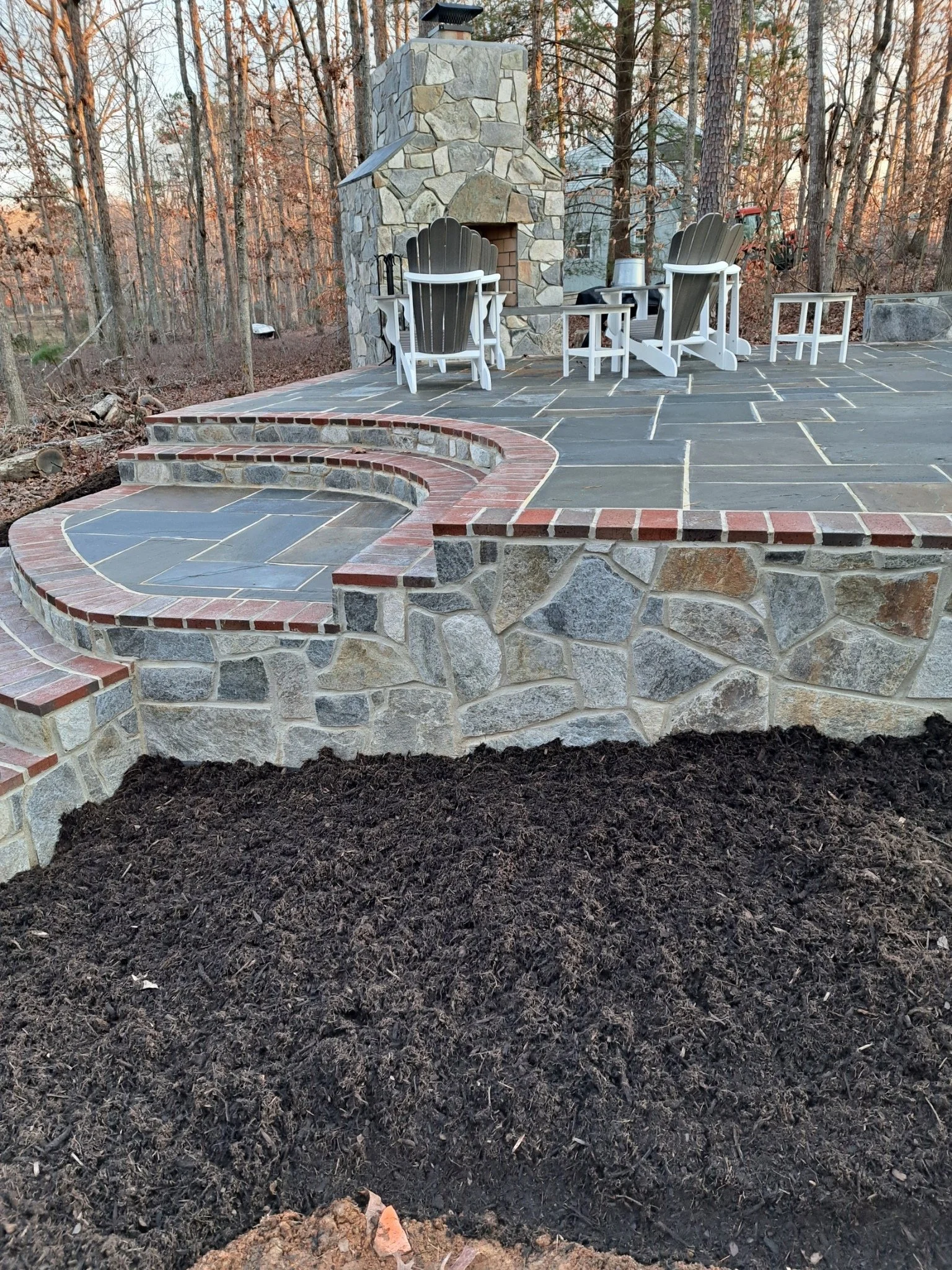 Backyard patio with stone fireplace, Adirondack chairs, small tables, and a garden bed with dark soil.