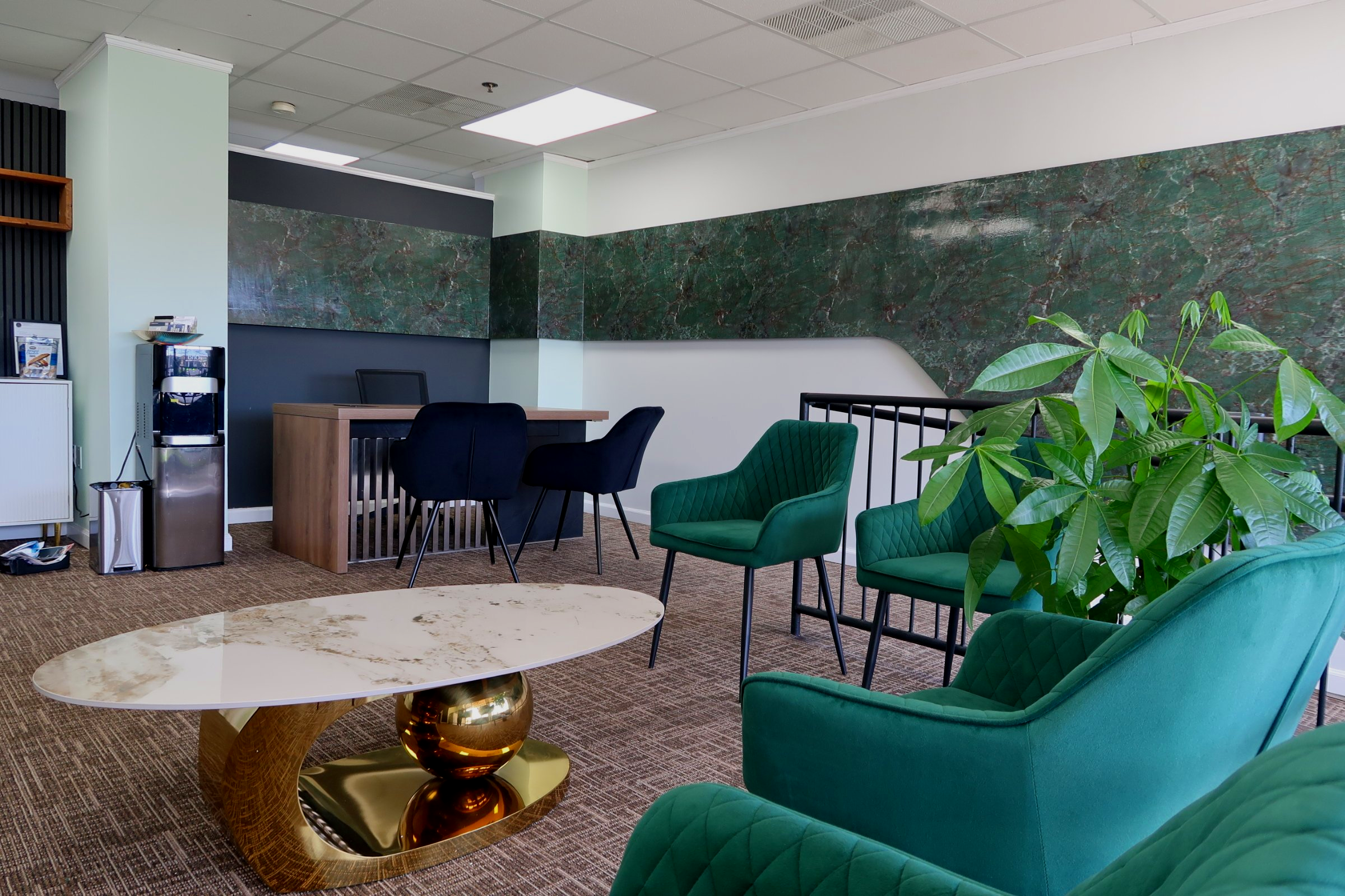 Modern office lounge with green armchairs, a white marble coffee table with a gold base, a large green plant, a small desk with two black chairs, and a wall featuring green marble accents.