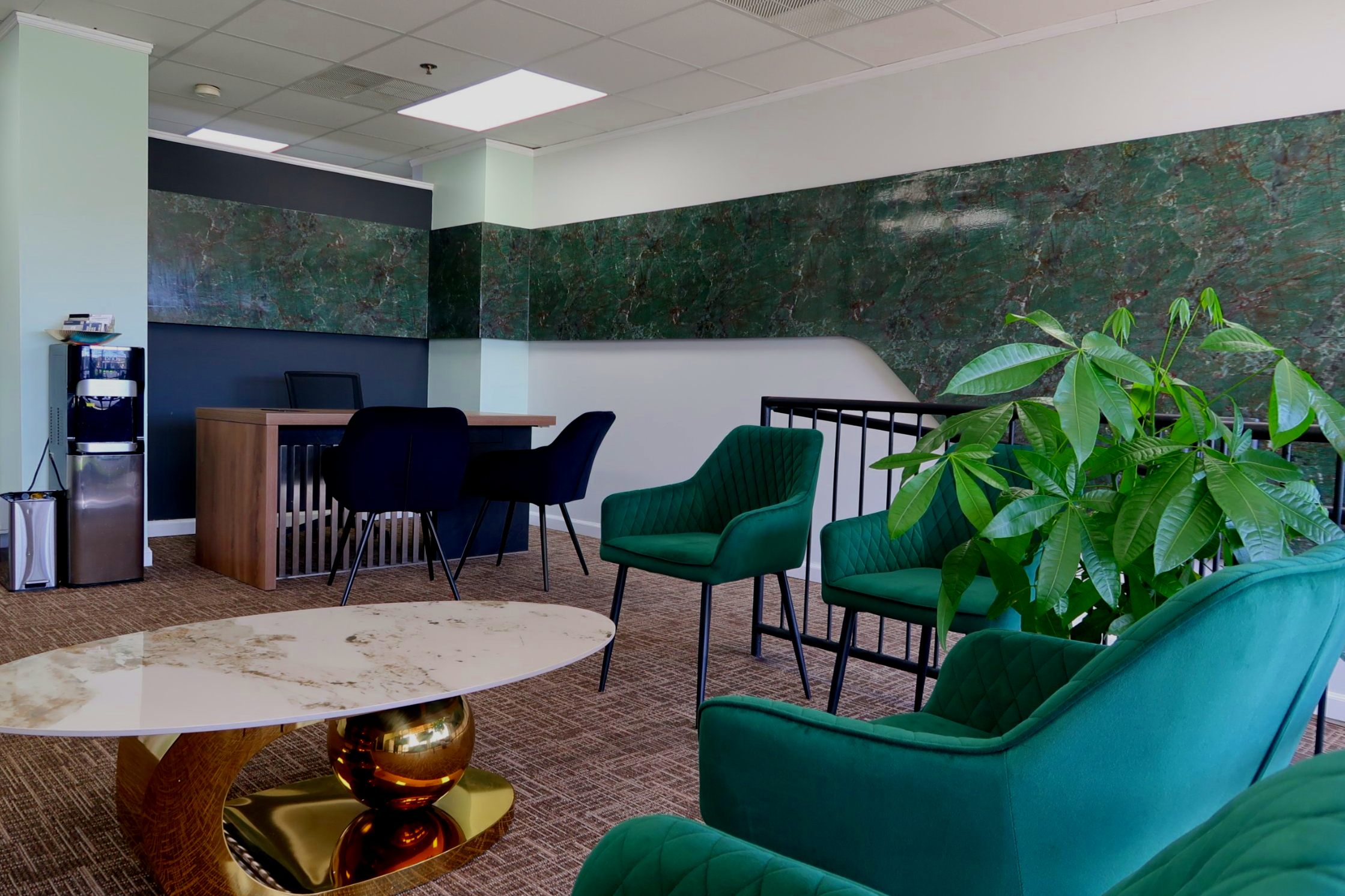 Modern office lounge with green armchairs, a white marble coffee table with a gold base, a large green plant, a small desk with two black chairs, and a wall featuring green marble accents.