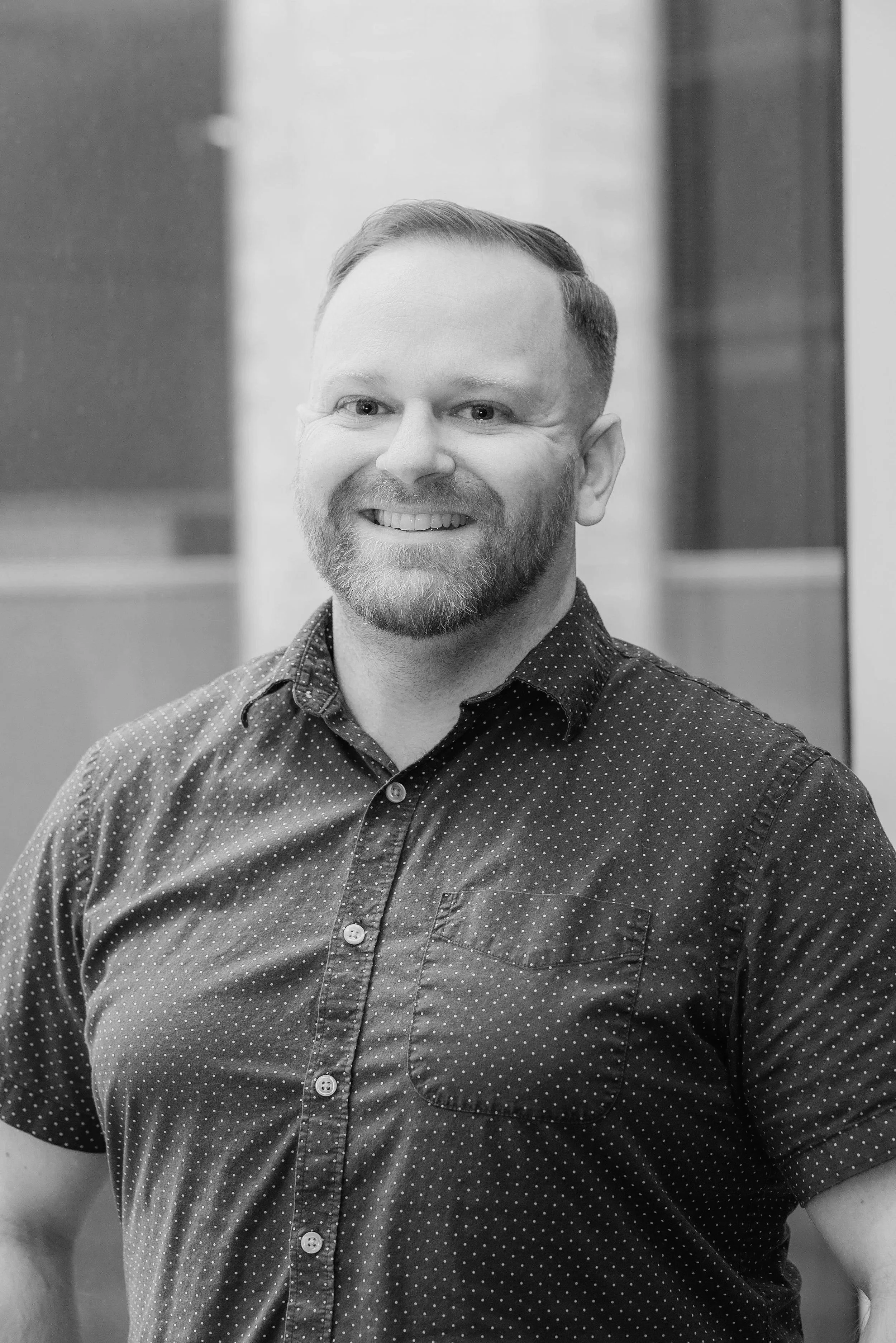 Black and white portrait of a smiling woman with medium-length hair and a necklace, standing against a plain dark background.