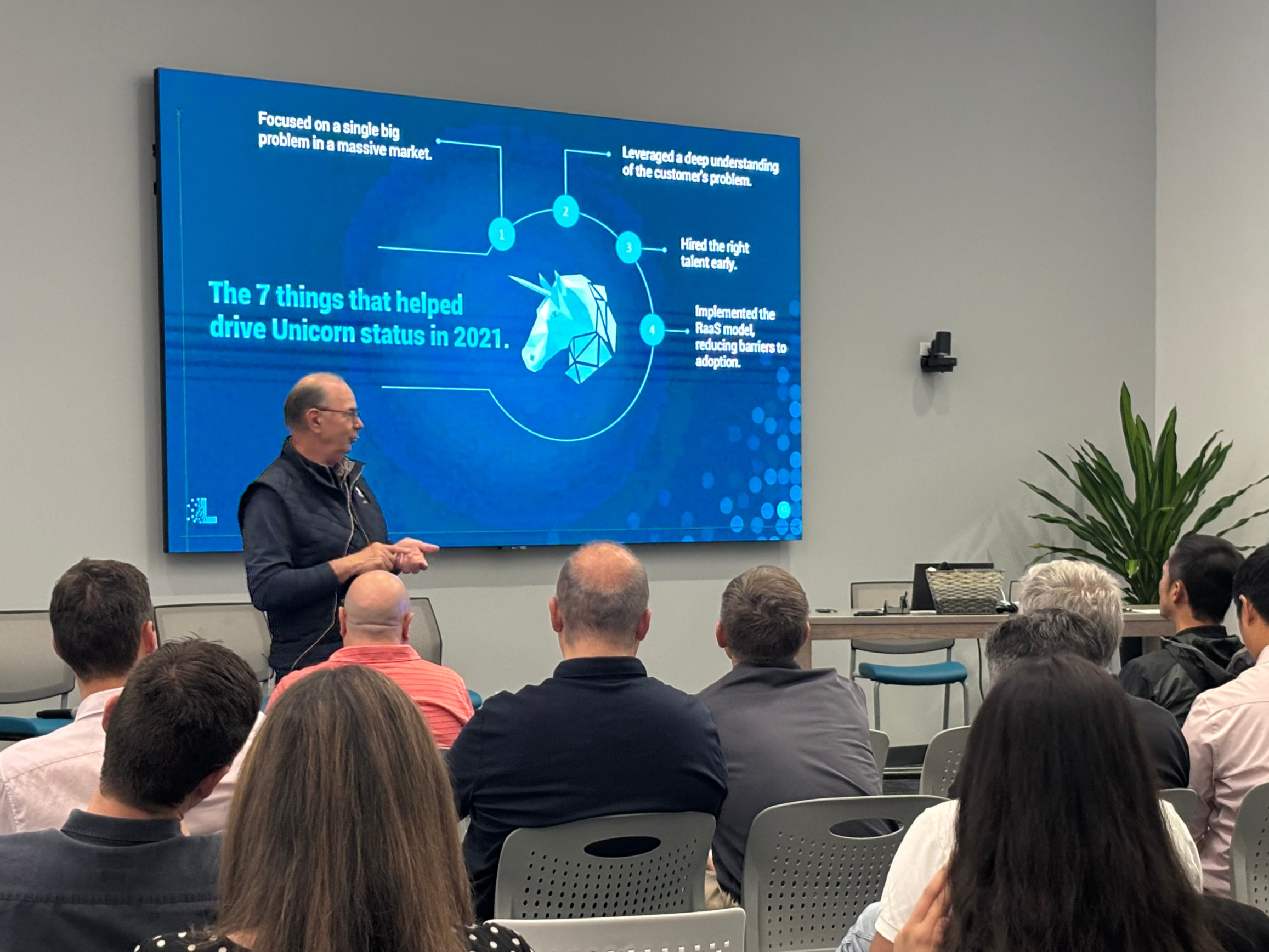 Rick giving a presentation to an audience in a conference room. The slideshow on the large screen behind him displays the title, "The 7 things that helped drive Unicorn status in 2021," with a graphic of a unicorn head and numbered points around it.