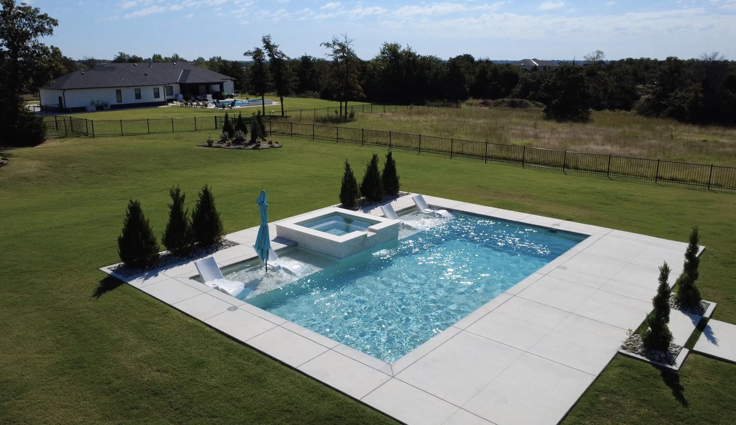 A backyard swimming pool with whirlpool, surrounded by green grass and small trees, with a house and trees in the background under a blue sky.