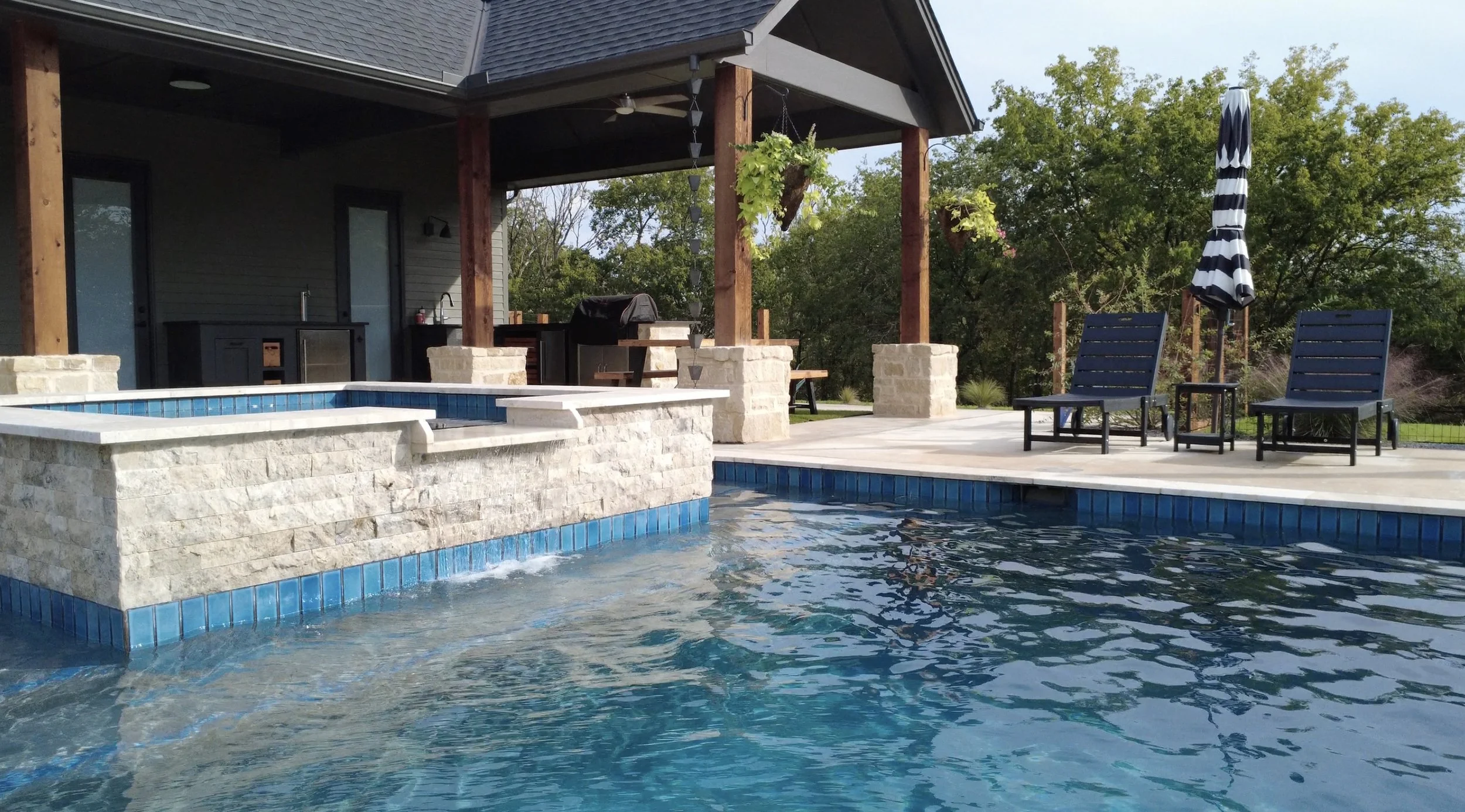 Swimming pool with a hot tub, two lounge chairs, and a striped umbrella on a patio with trees in the background.
