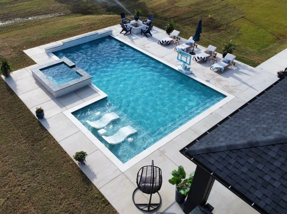 Aerial view of a modern backyard with a rectangular swimming pool, hot tub, poolside lounge chairs with striped towels, a seating area with patio furniture, potted plants, and a grassy lawn surrounding the area.