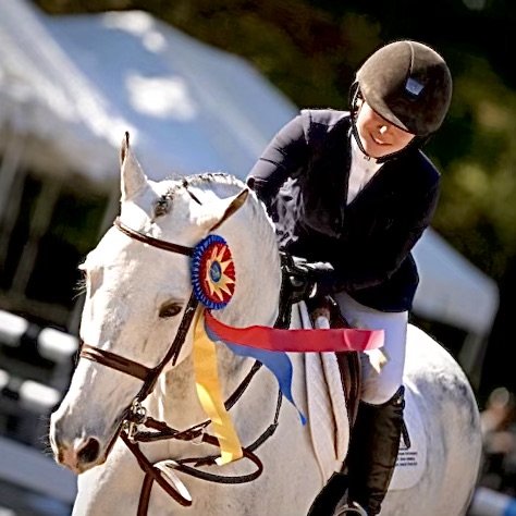 A smiling woman wearing a helmet and a black riding jacket riding a white horse with a ribbon on its bridle, during a horse riding competition.