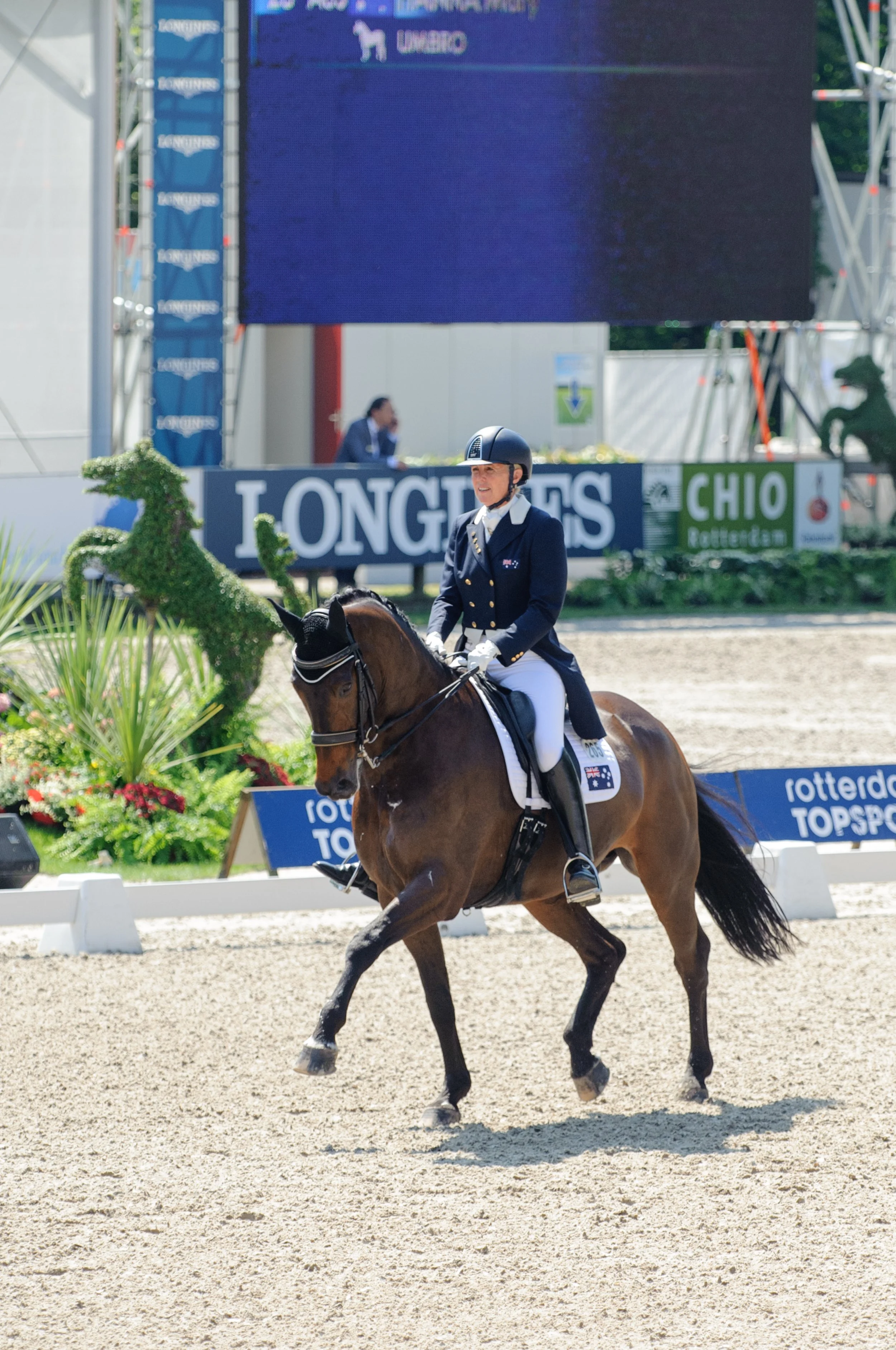 A horseback rider in formal equestrian attire riding a brown horse at a competitive event, with a large screen in the background displaying the Longines logo and other sponsors, and decorative plants along the riding arena.