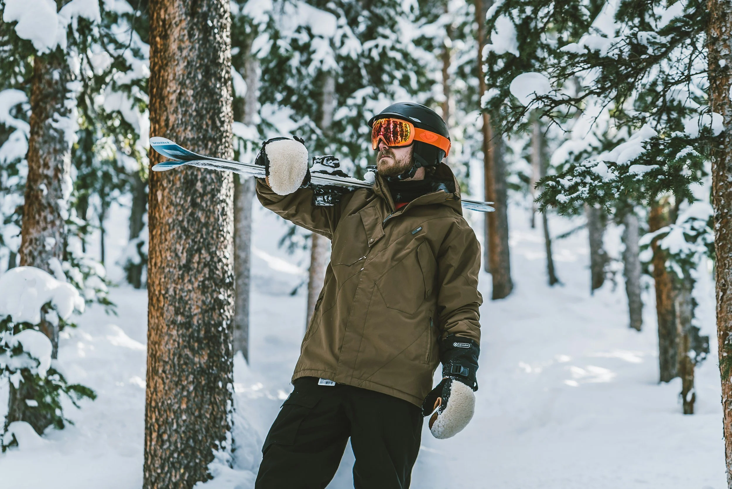 Big bear Skier in snowy forest carrying skis, wearing brown jacket, helmet, goggles, and gloves.