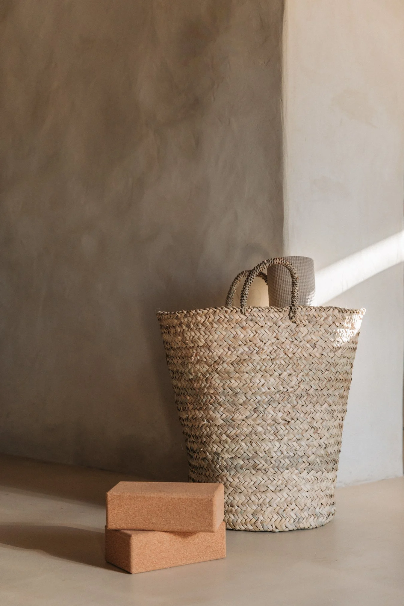 A woven straw tote bag with handles and a rolled-up towel inside, placed on the floor beside two stacked terracotta-colored bricks, with sunlight casting shadows in the background.