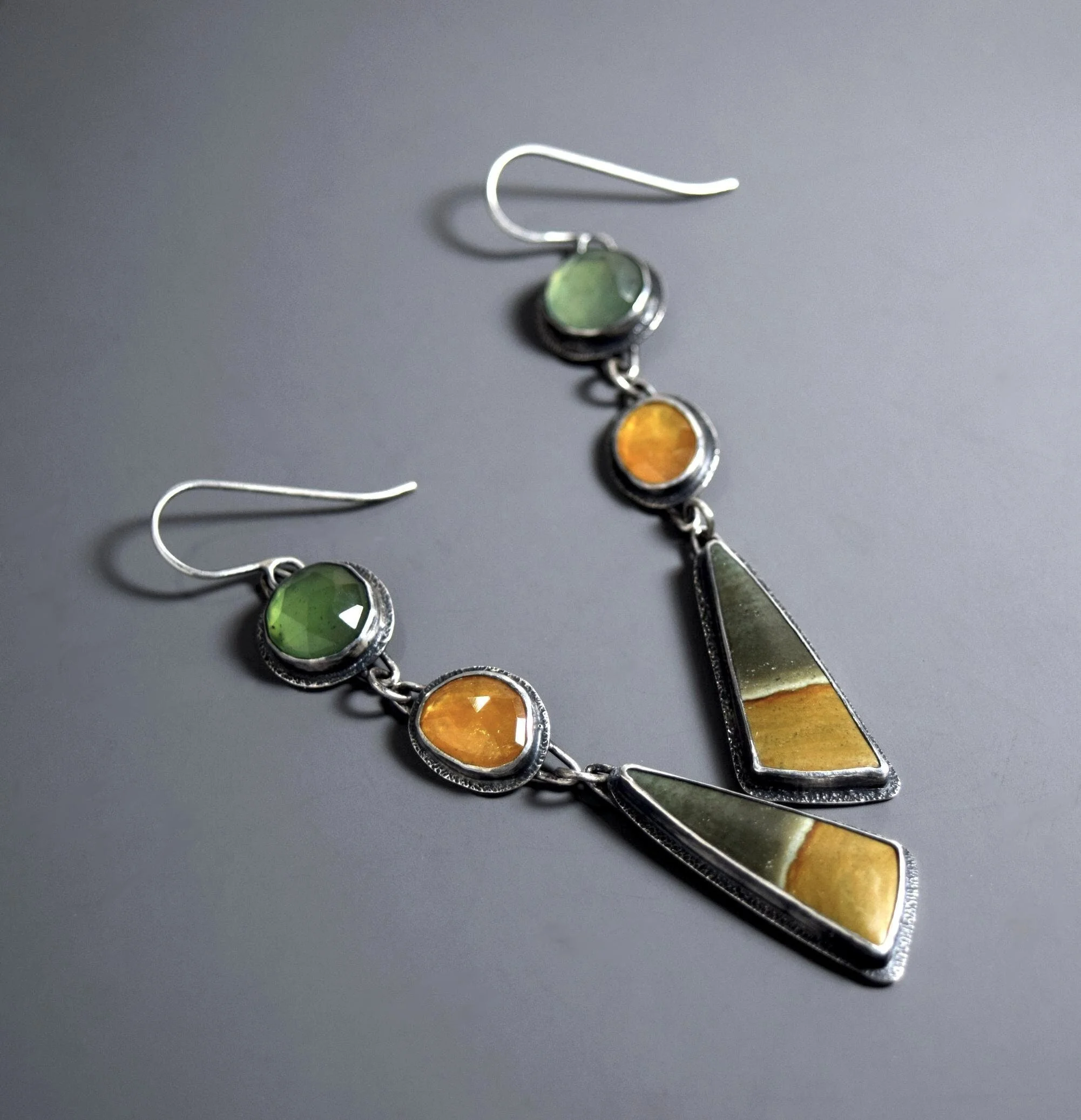 Pair of silver dangle earrings with colorful oval and round stones in green, orange, and yellowish tones, featuring geometric shapes and designs.