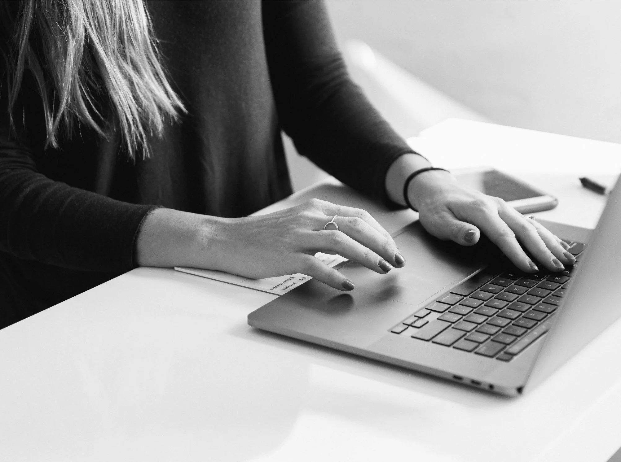 A woman typing on a laptop at a white table with a smartphone and a notepad nearby.