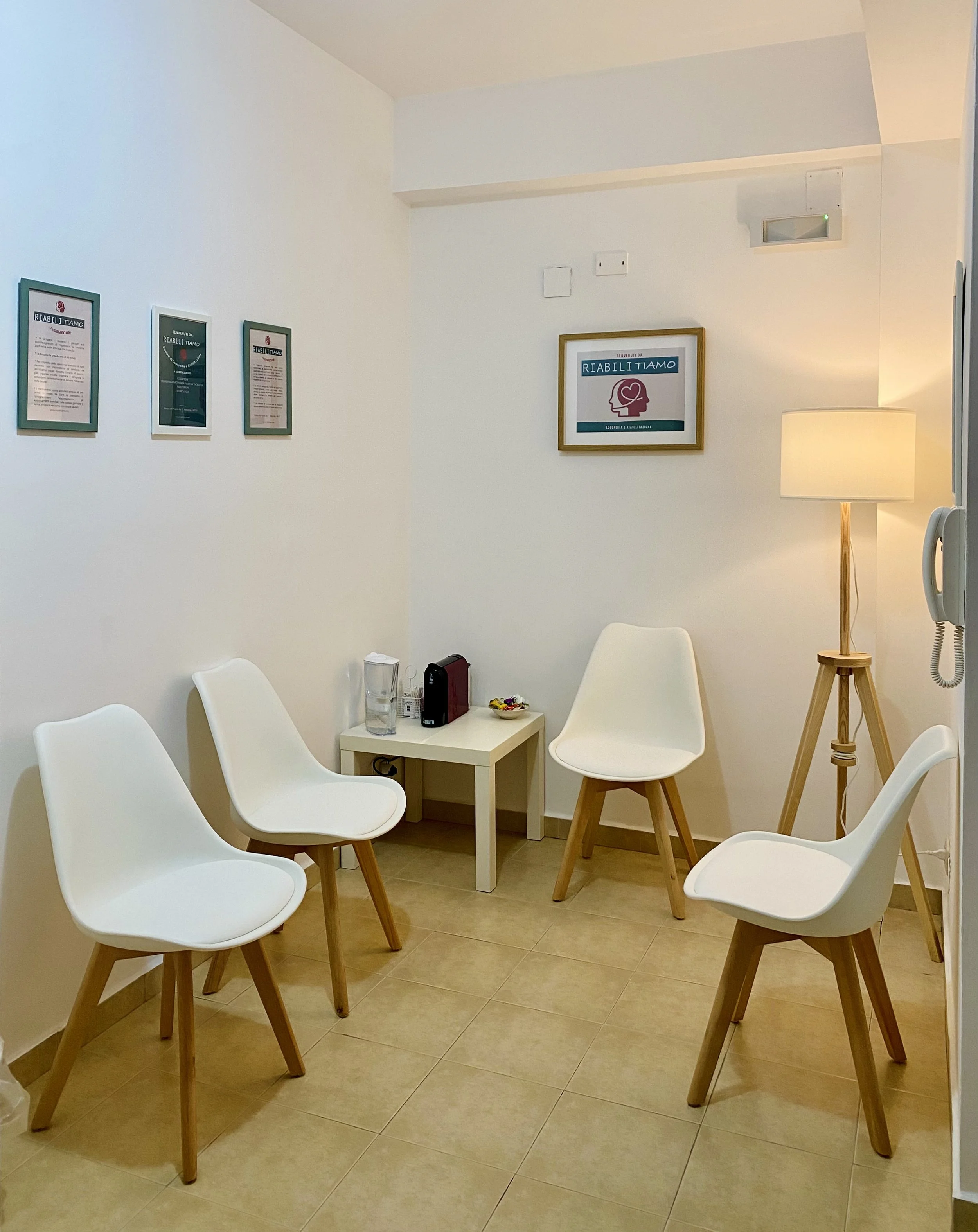 Sala di attesa con sedie bianche in stile Scandinavo disposte intorno a un tavolino con bicchieri, una scatola e caramelle. Parallele ai mobili, una lampada da terra con abat-jour e un quadro con testo e logo. Pareti bianche con cornice dorata. Muro 