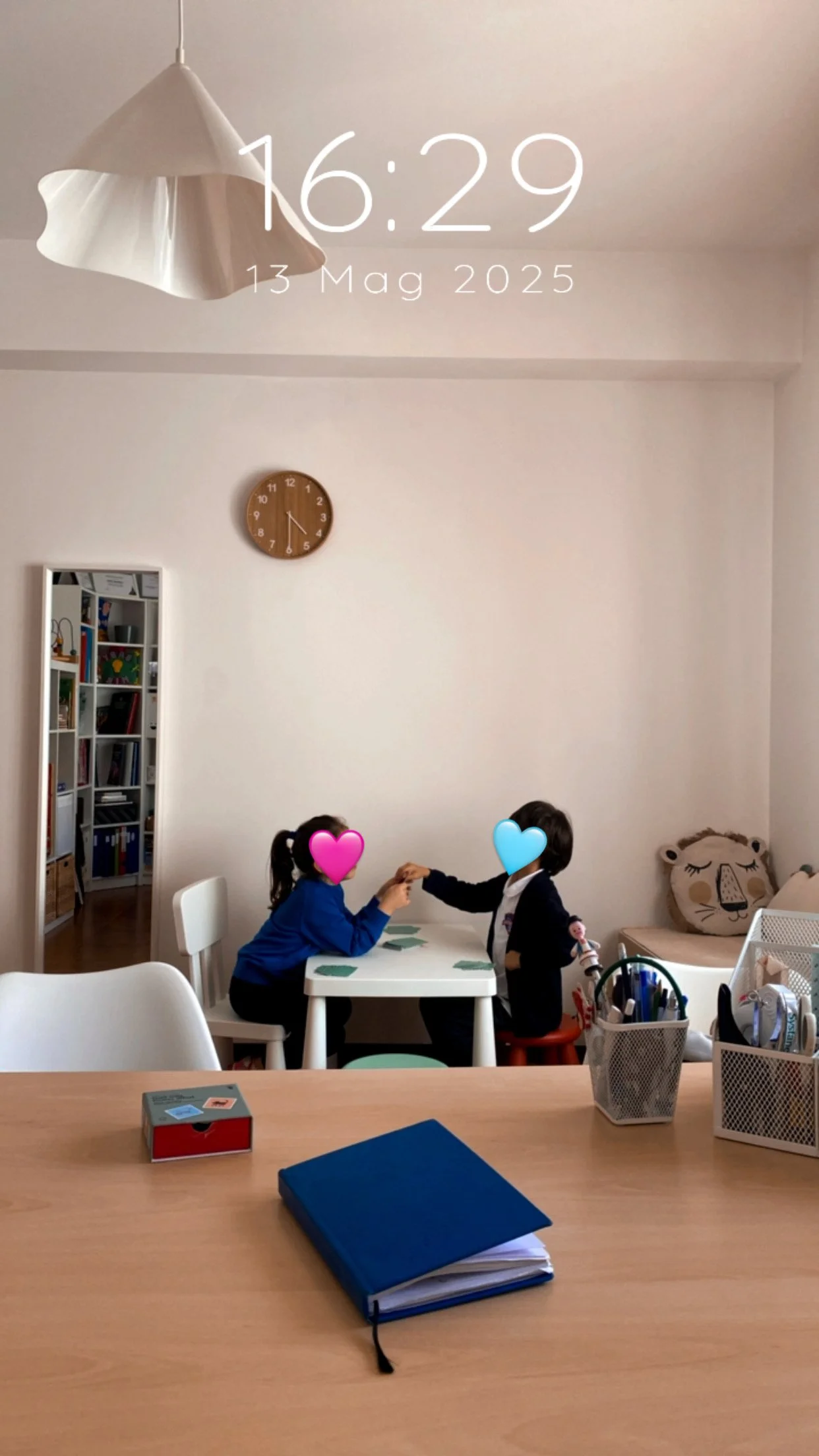 Due bambini seduti a un tavolo che si tengono per le mani in una stanza luminosa, con lo sfondo di una parete bianca, un orologio e un divano con cuscini decorativi a forma di faccia di animale.