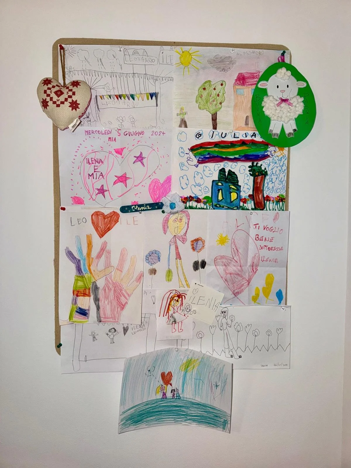 Murale con disegni fatti da bambini, tra cui case, alberi, arcobaleni, cuori, personaggi stilizzati, e scritte in italiano, decorata con cuori di stoffa e un cartone con una pecora di cotone.
