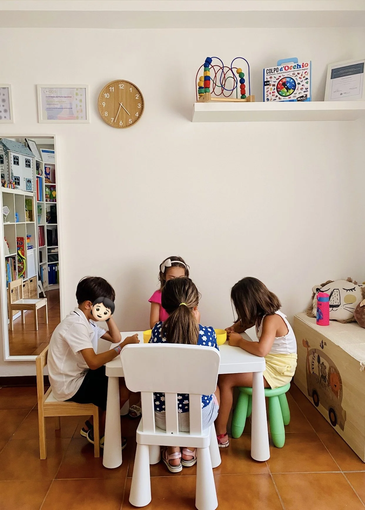 Cinque bambini seduti attorno a un tavolo bianco che giocano, in una stanza con scaffali pieni di libri e giochi, decorazioni infantili e un orologio a parete. Una finestra mostra una stanza con mobili simili.