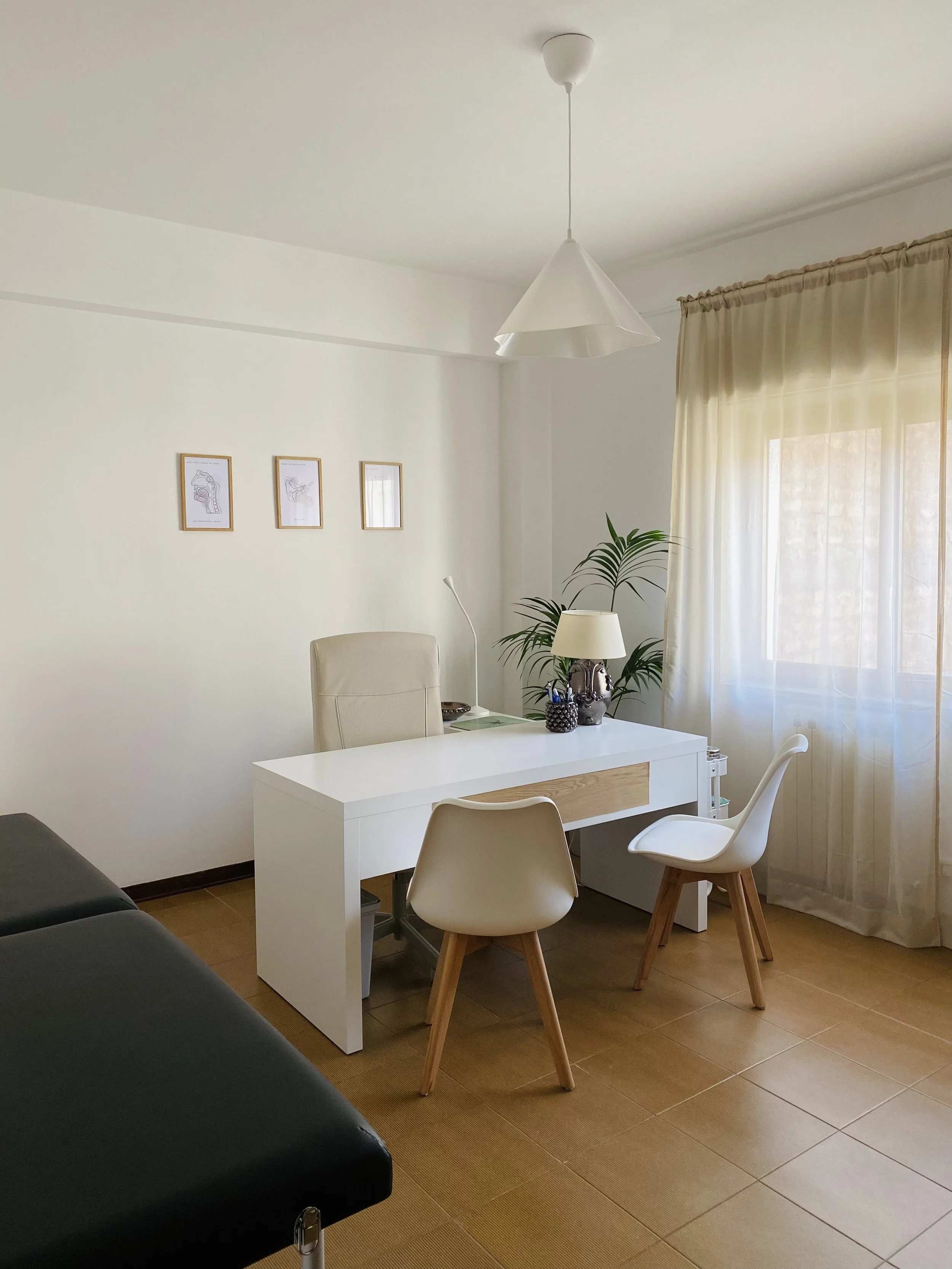 Studio con un tavolo bianco, una sedia con gambe di legno, una lampada, un vaso con piante e una finestra con tende beige.