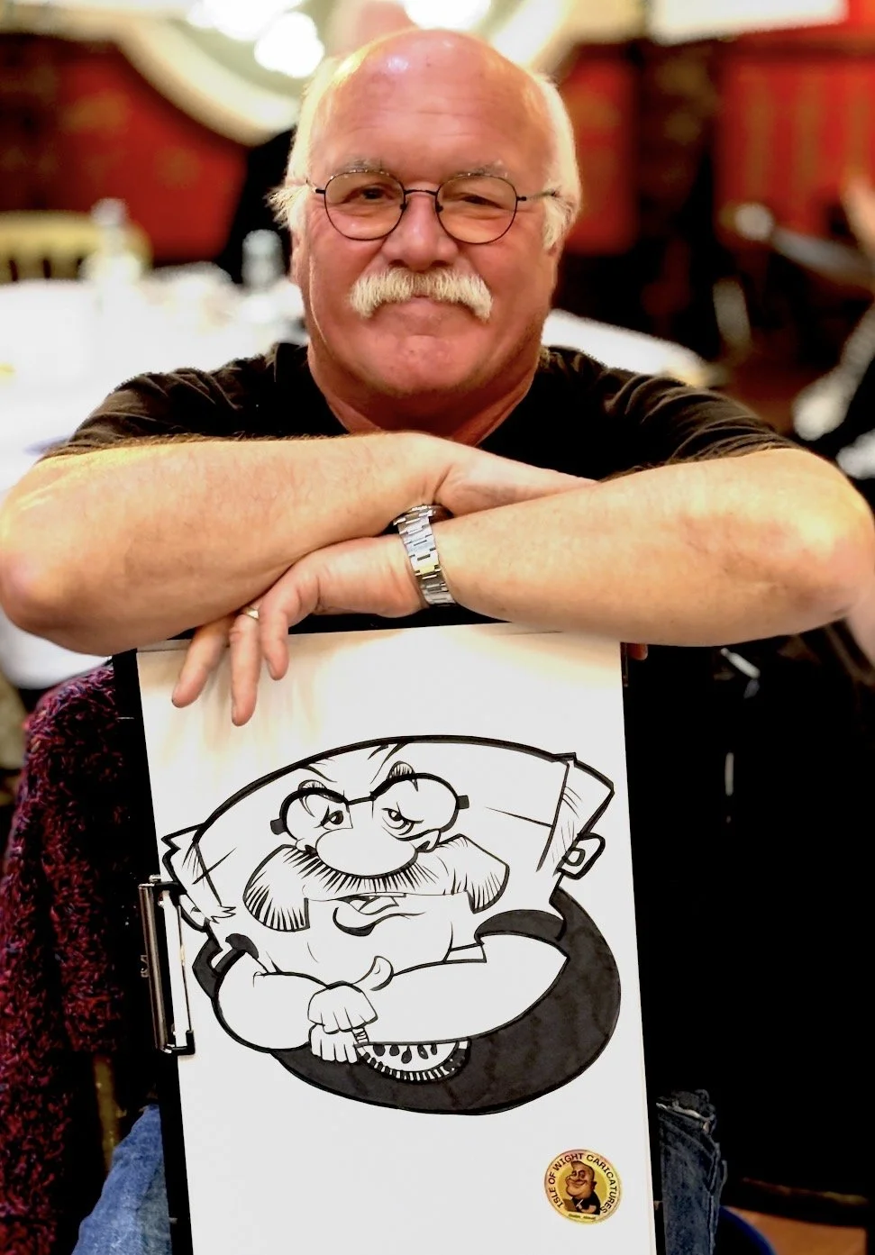 A smiling elderly man with glasses and a mustache, sitting at a table, resting his arms on a drawing of a cartoon character with a beard and glasses, at an indoor event.