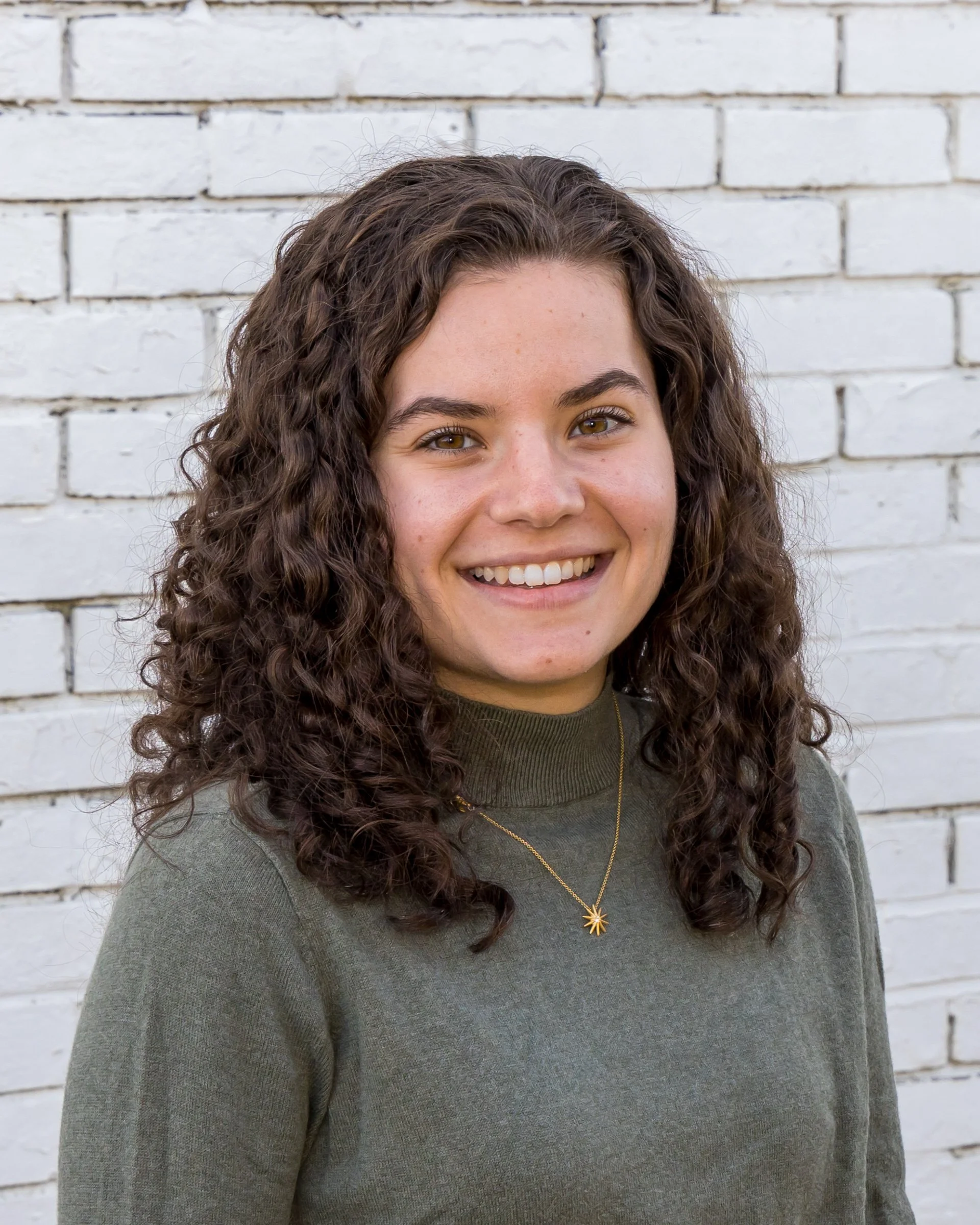 A woman with curly brown hair and light skin smiling, wearing a green turtleneck sweater and a gold necklace with a star-shaped pendant, standing in front of a white brick wall.