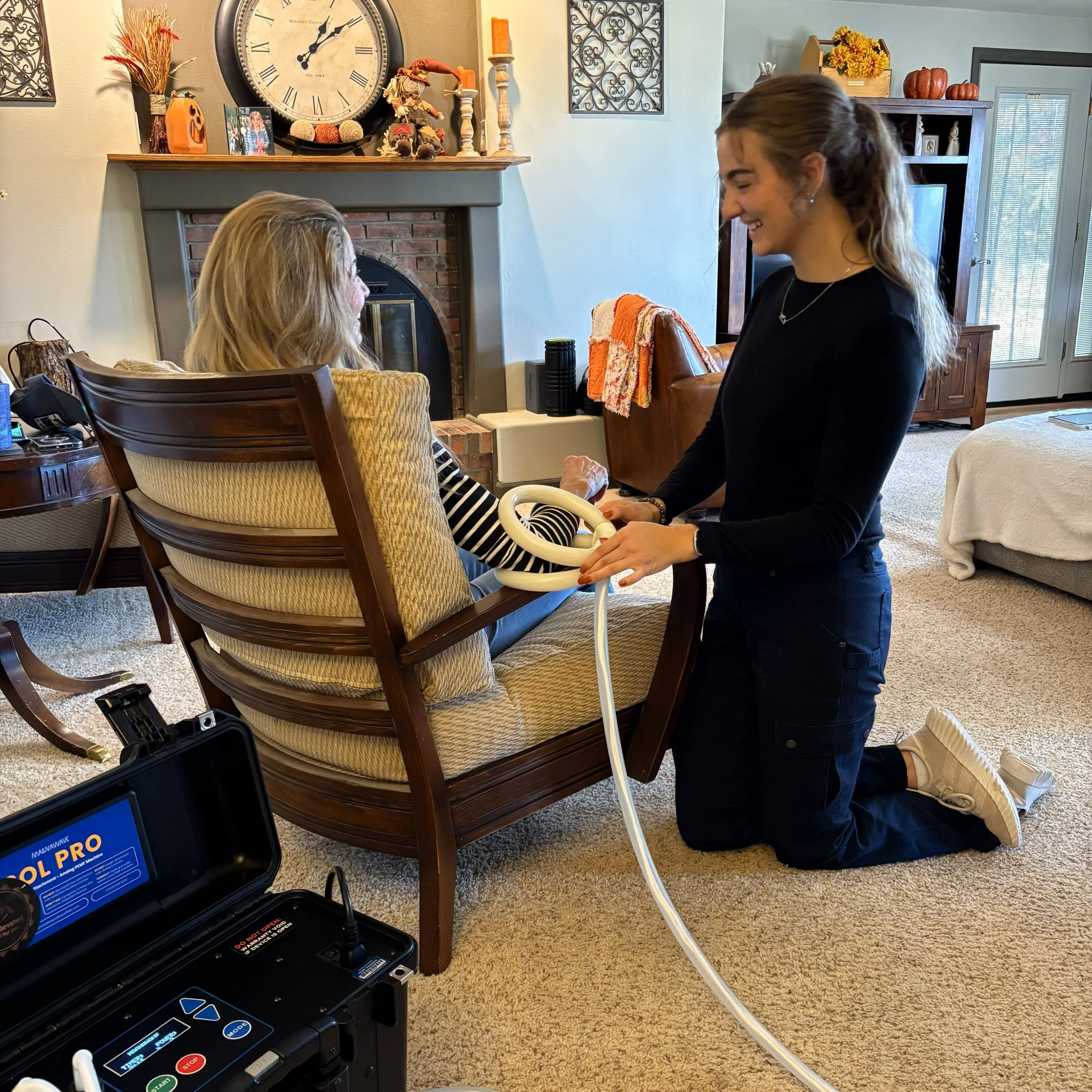 A young woman kneeling next to an elderly woman sitting in a wooden chair, with a pemf machine and pemf coil, smiling and engaging in conversation, in a warm living room decorated with fall themed items and a large clock above a fireplace.
