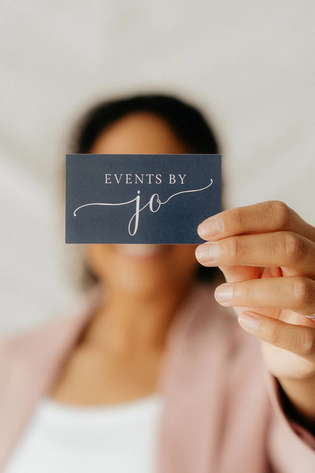 Person holding a navy business card that reads 'Events by Jo', with their face out of focus in the background.