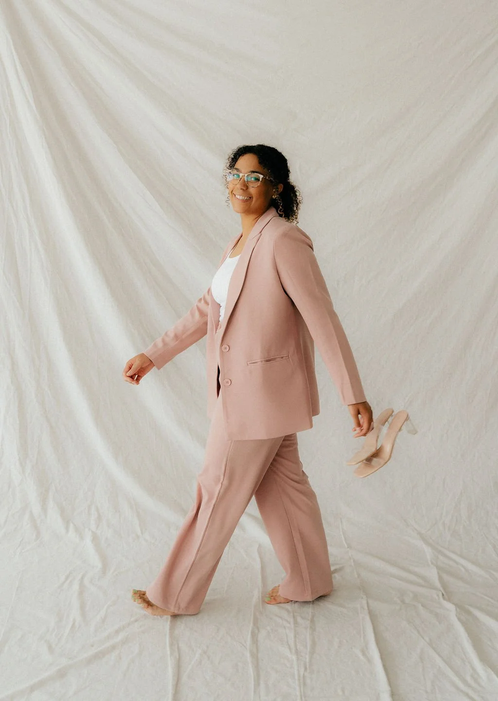 A woman in a pink suit walking barefoot with beige high heels in her hand, smiling, against a light fabric backdrop.