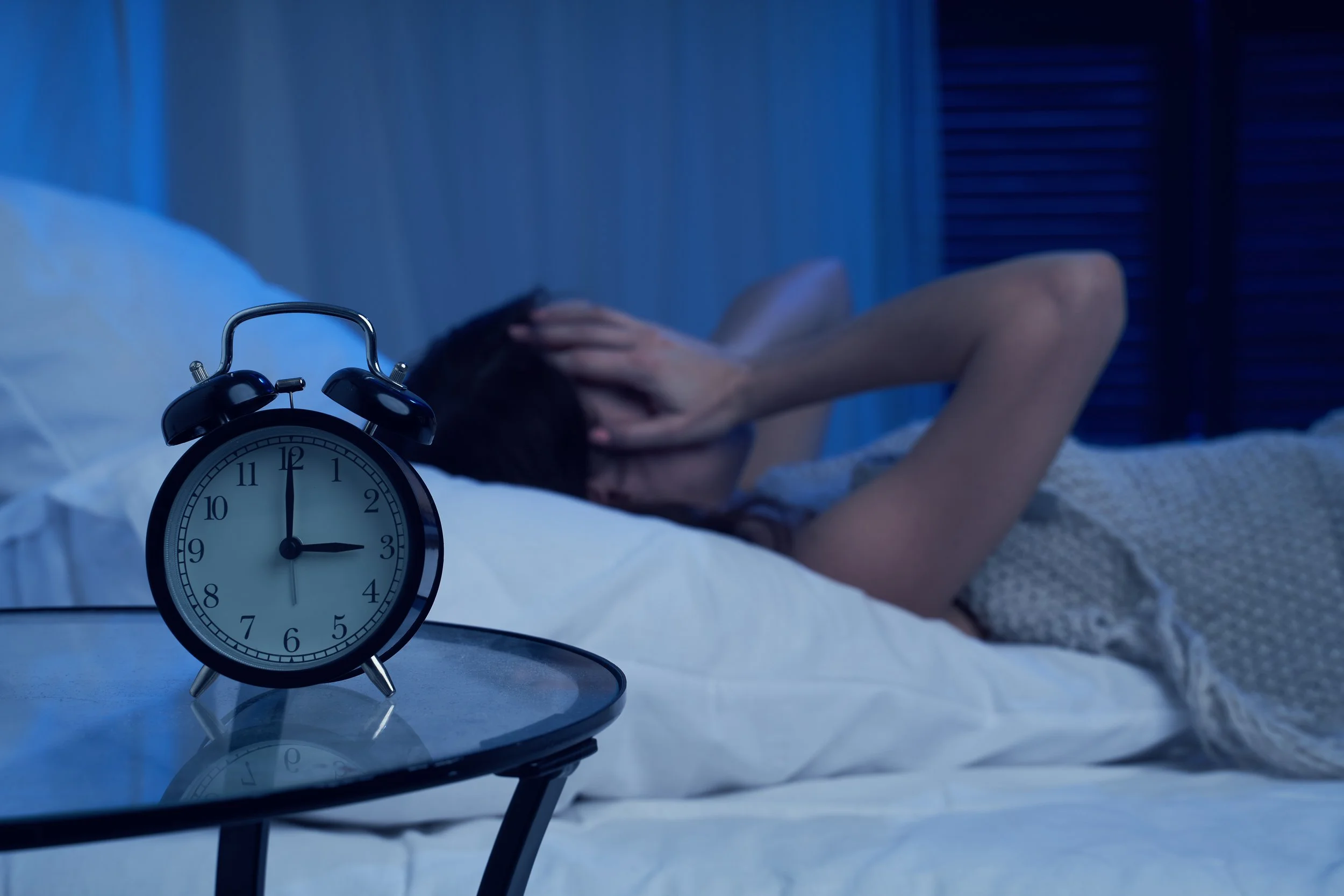 Alarm clock showing 3:00 AM on bedside table in front of a person lying in bed, holding their head with one hand, appearing distressed.