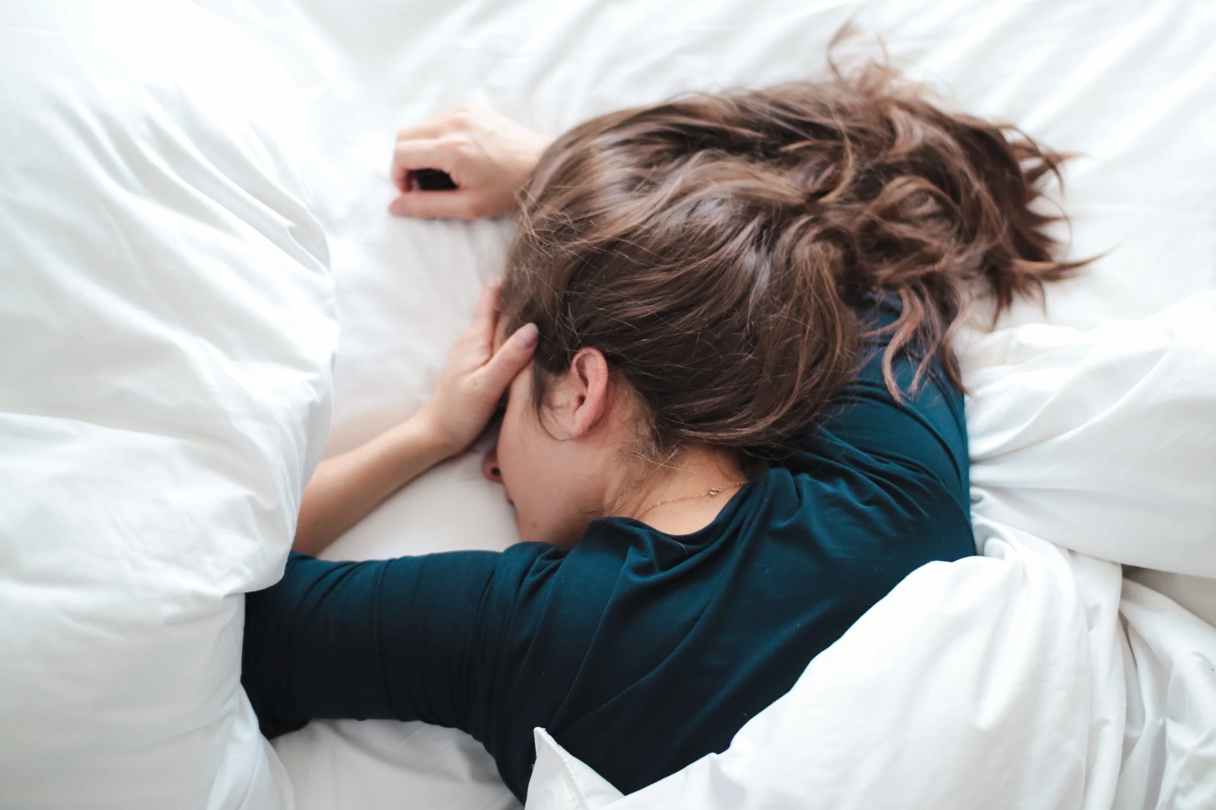 A woman with long brown hair sleeping on a bed with white sheets, lying on her side with her hand resting on her head.