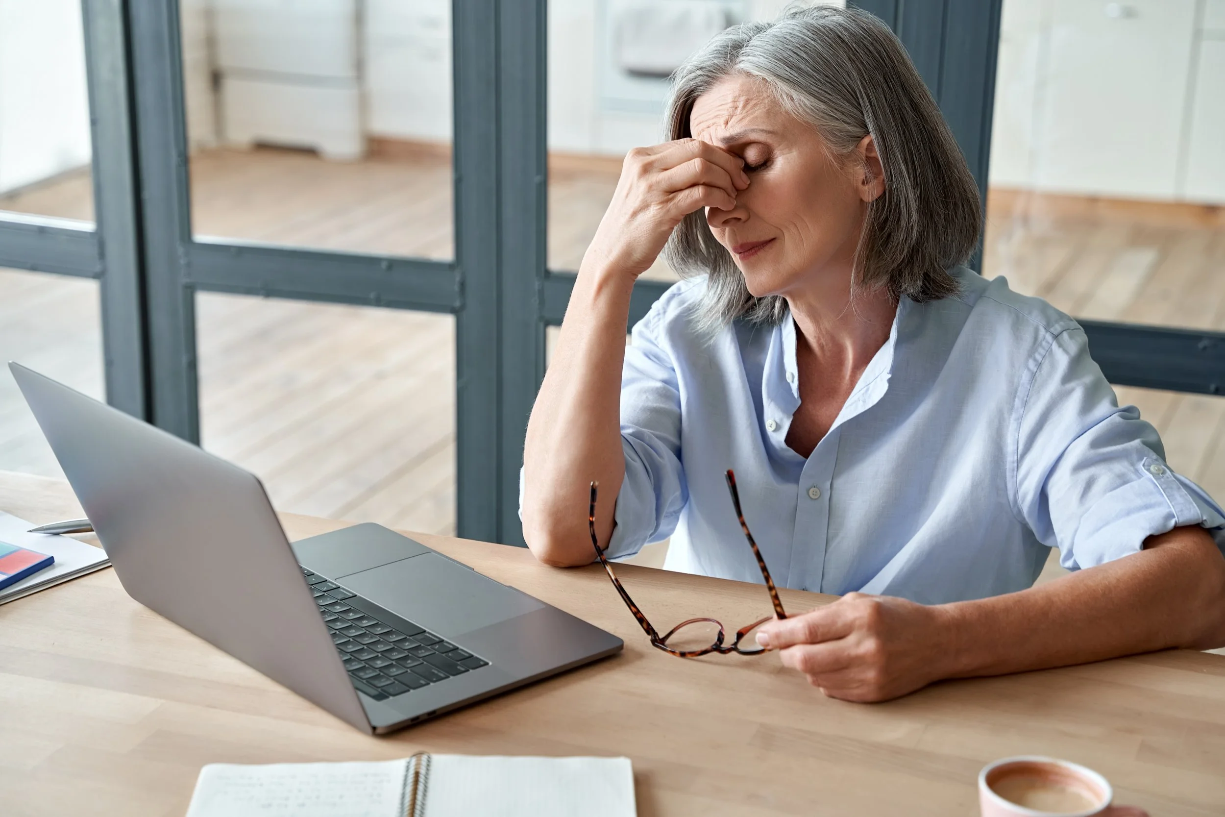 An older woman sitting at a wooden table with a laptop, holding her glasses and pinching the bridge of her nose, appearing stressed or tired.