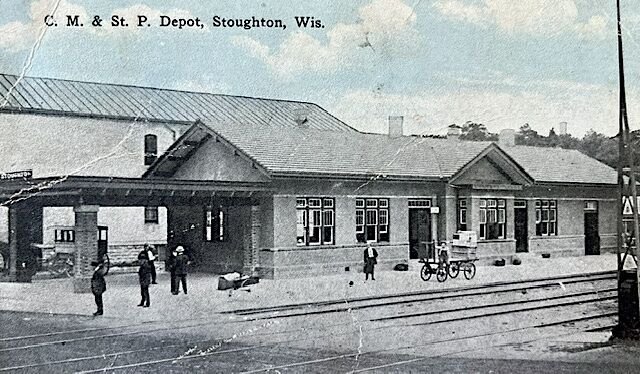 Historic postcard showing Stoughton’s passenger depot, with people on the platform and railroad tracks in the foreground.