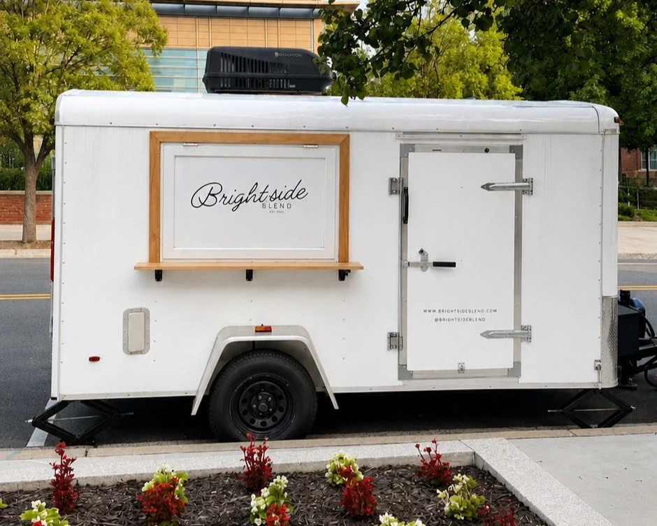 A white food truck named 'Brightside Blend' parked on the street with a large window and a side door. The truck has a small shelf and is surrounded by greenery and flowers.