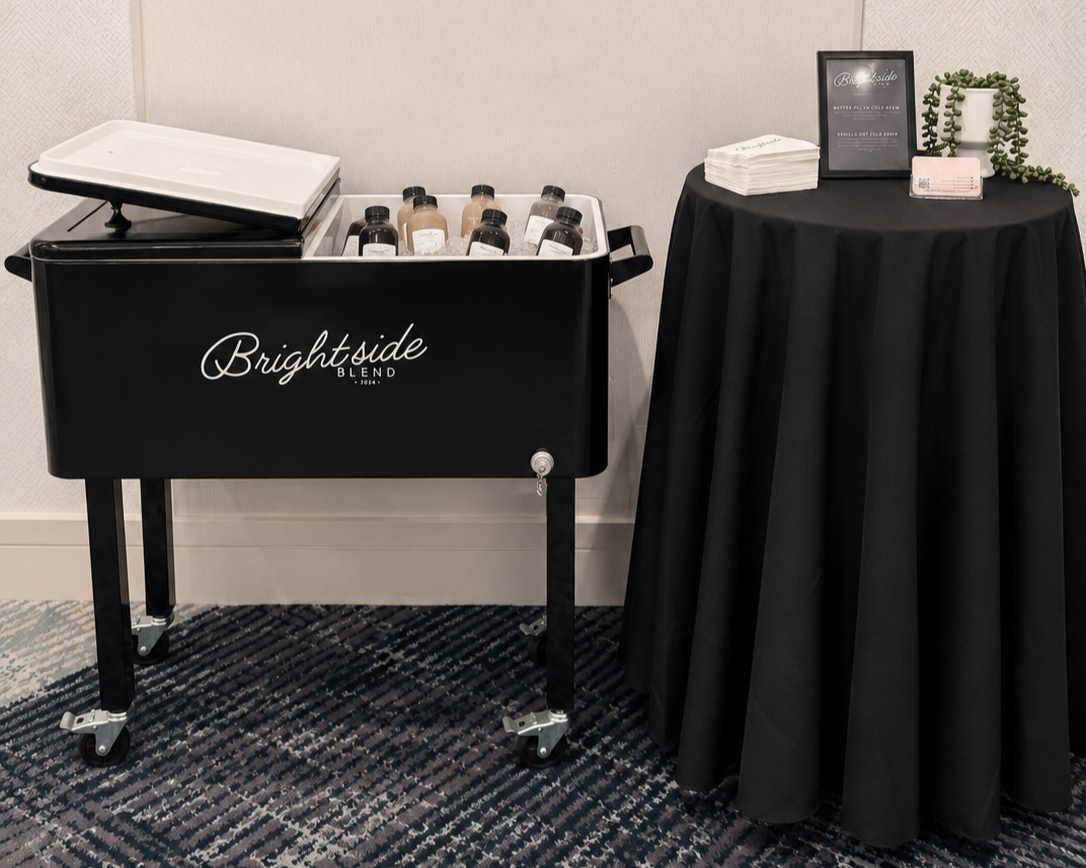 A black rolling cart labeled 'Brightside Blend' filled with bottles of bottled beverages, next to a black table covered with a cloth, holding a framed sign, neatly folded napkins, a small potted plant, and a box of tea bags.