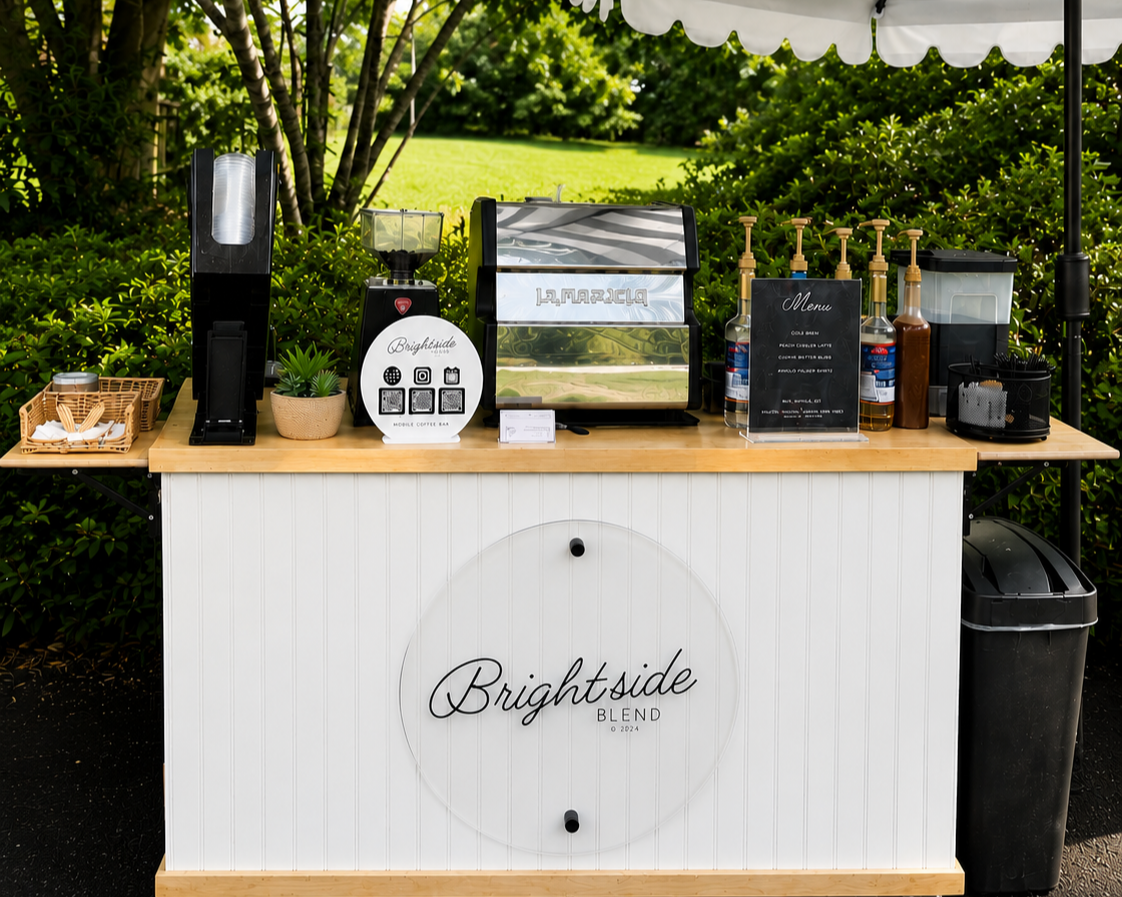 Outdoor coffee station setup with a La Marzocco espresso machine, a grinder, condiments, and menu signs under a white canopy, surrounded by greenery.