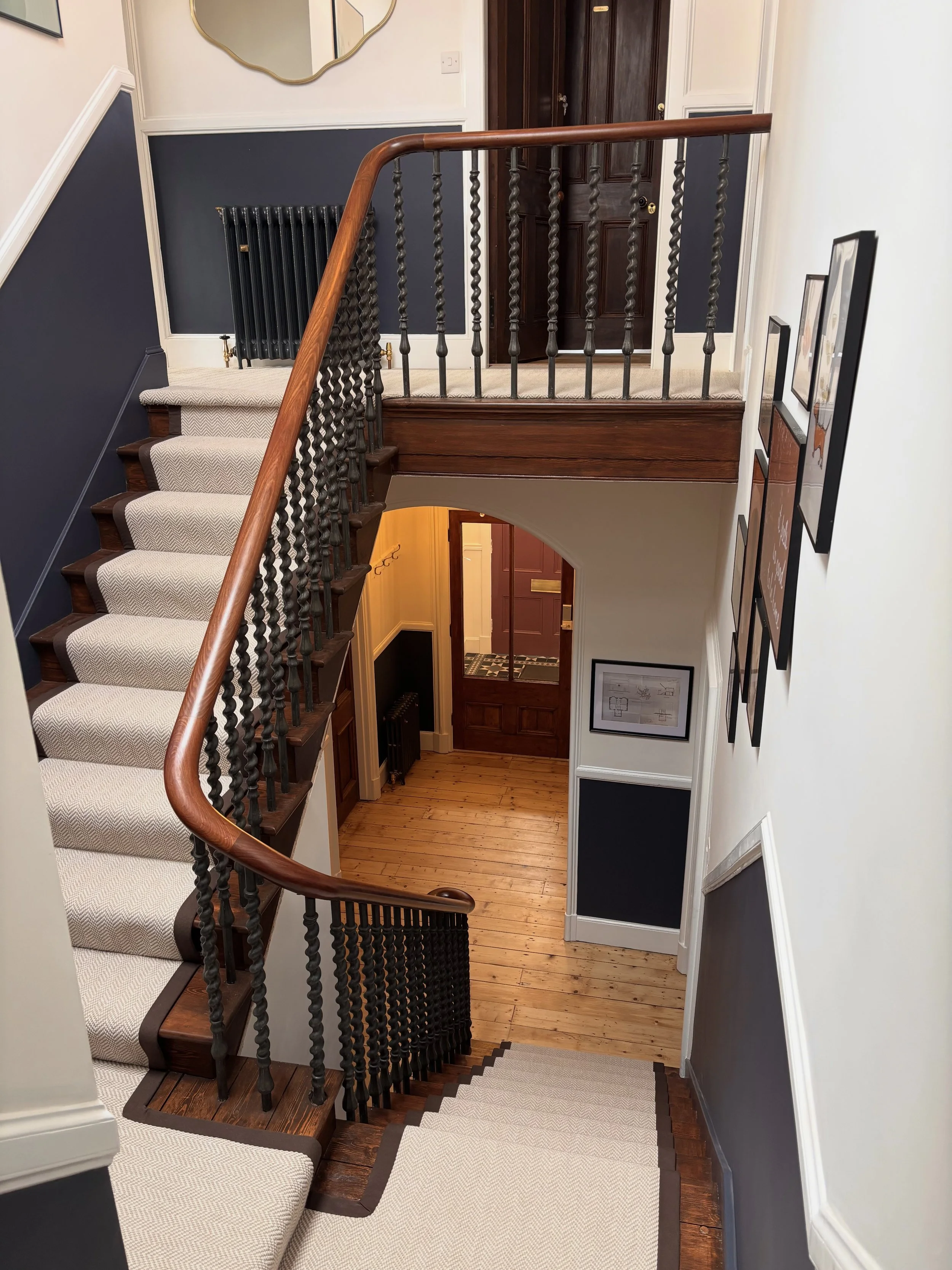 Interior view of a staircase with wooden handrails, beige carpeting, and dark blue and white walls leading to a ground floor foyer with a wooden floor, hallway, and framed pictures on the wall.