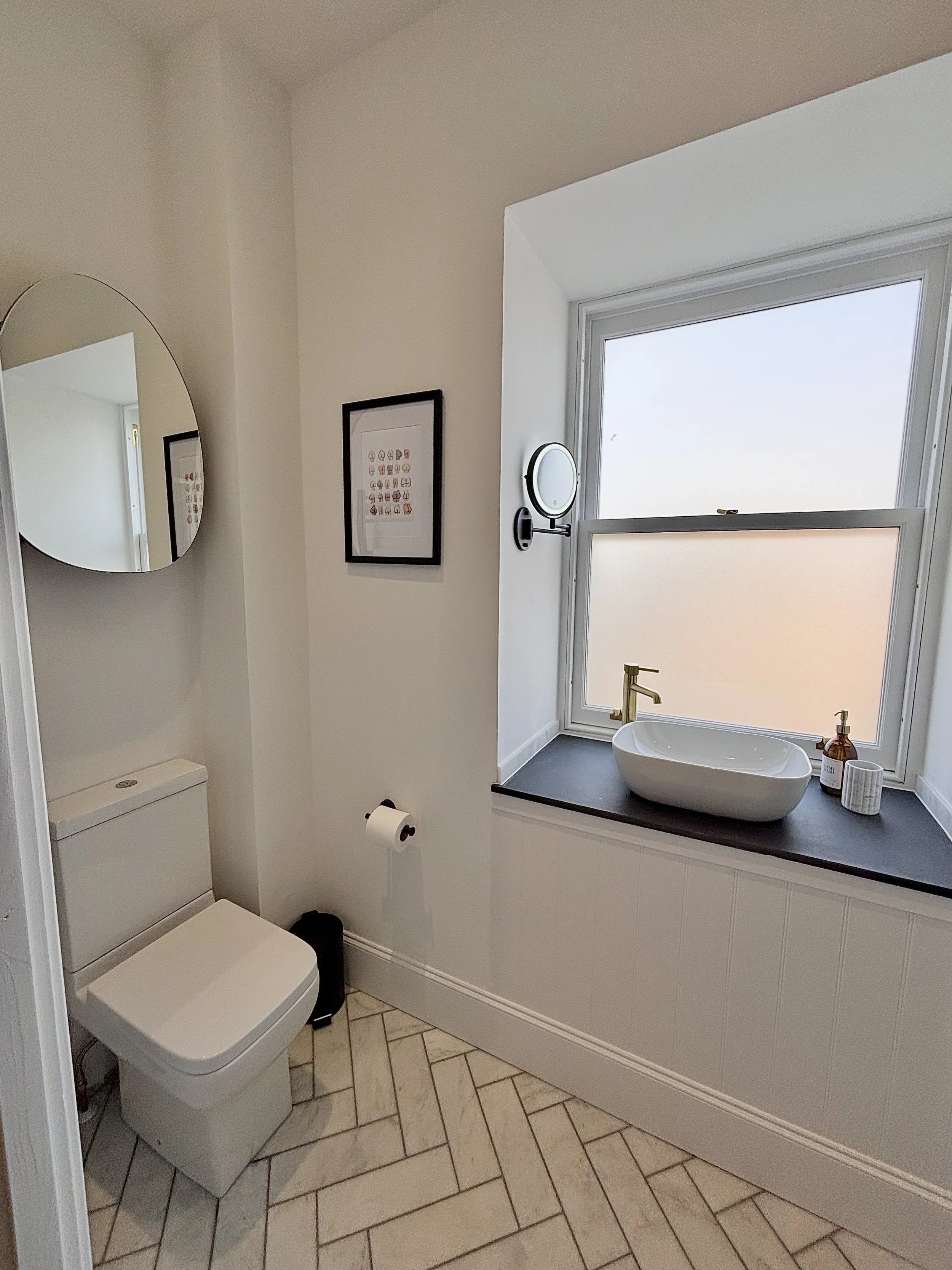 A modern bathroom with a white toilet, a black countertop with a white vessel sink, a brass faucet, a large window for natural light, a round mirror, a wall clock, framed artwork, and bathroom toiletries.