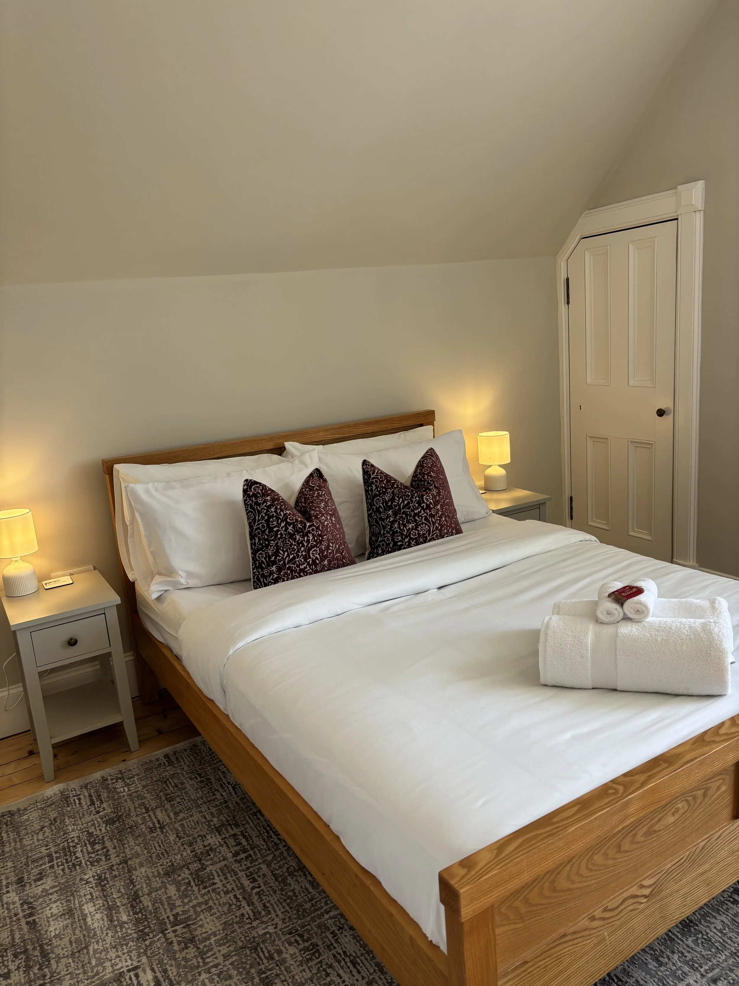 A neatly made bed with white sheets and several purple patterned pillows, flanked by two nightstands with lamps, set against a beige wall with a white door with molding.