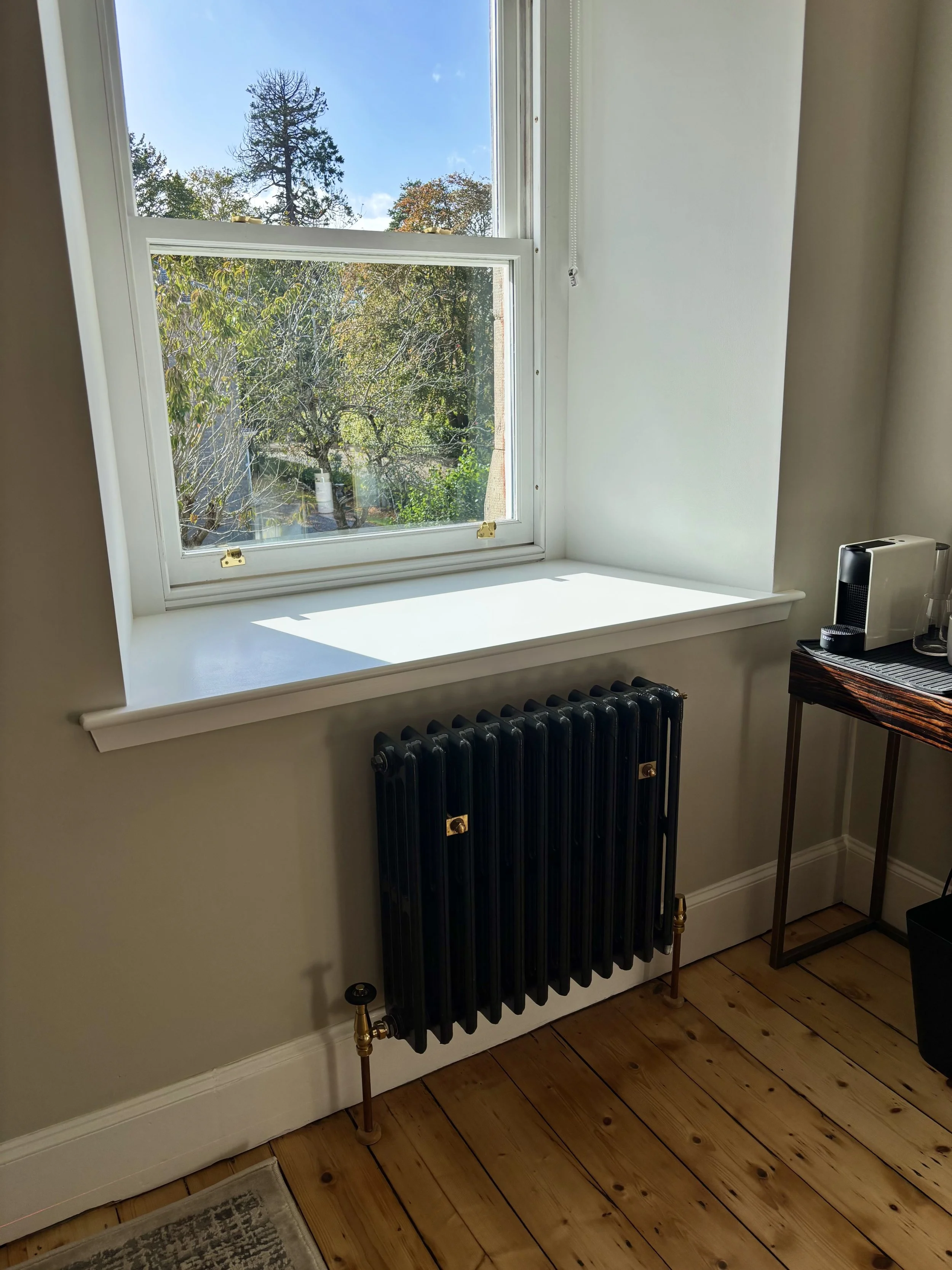 A window overlooking trees and a backyard, with a black radiator below the window and a dark wooden table to the right holding an air fryer and a kettle, on a wooden floor with a gray rug nearby.