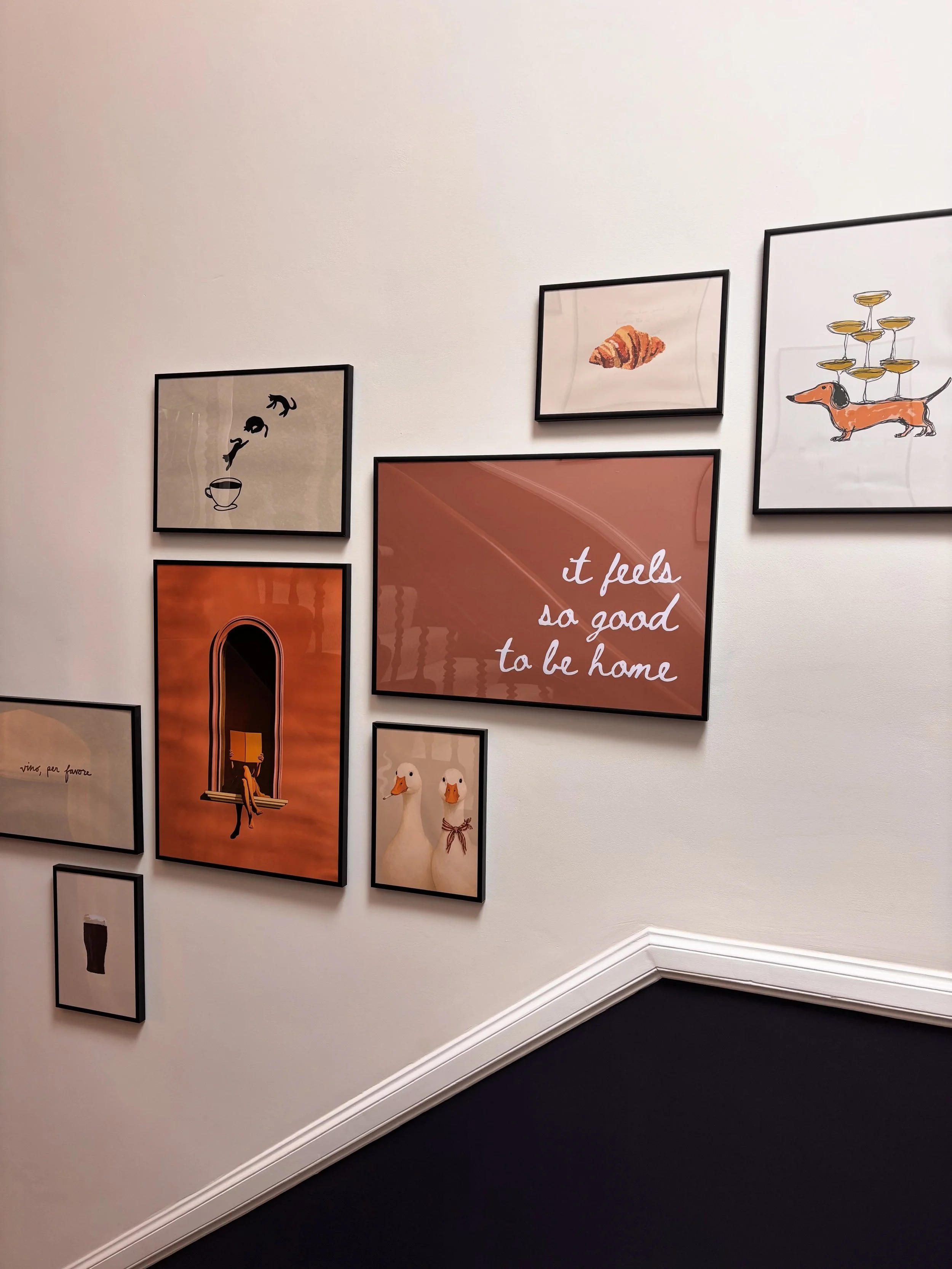 Gallery wall with various framed art pieces, including illustrations of animals, food, and motivational quotes, in a room with beige walls and dark carpet.