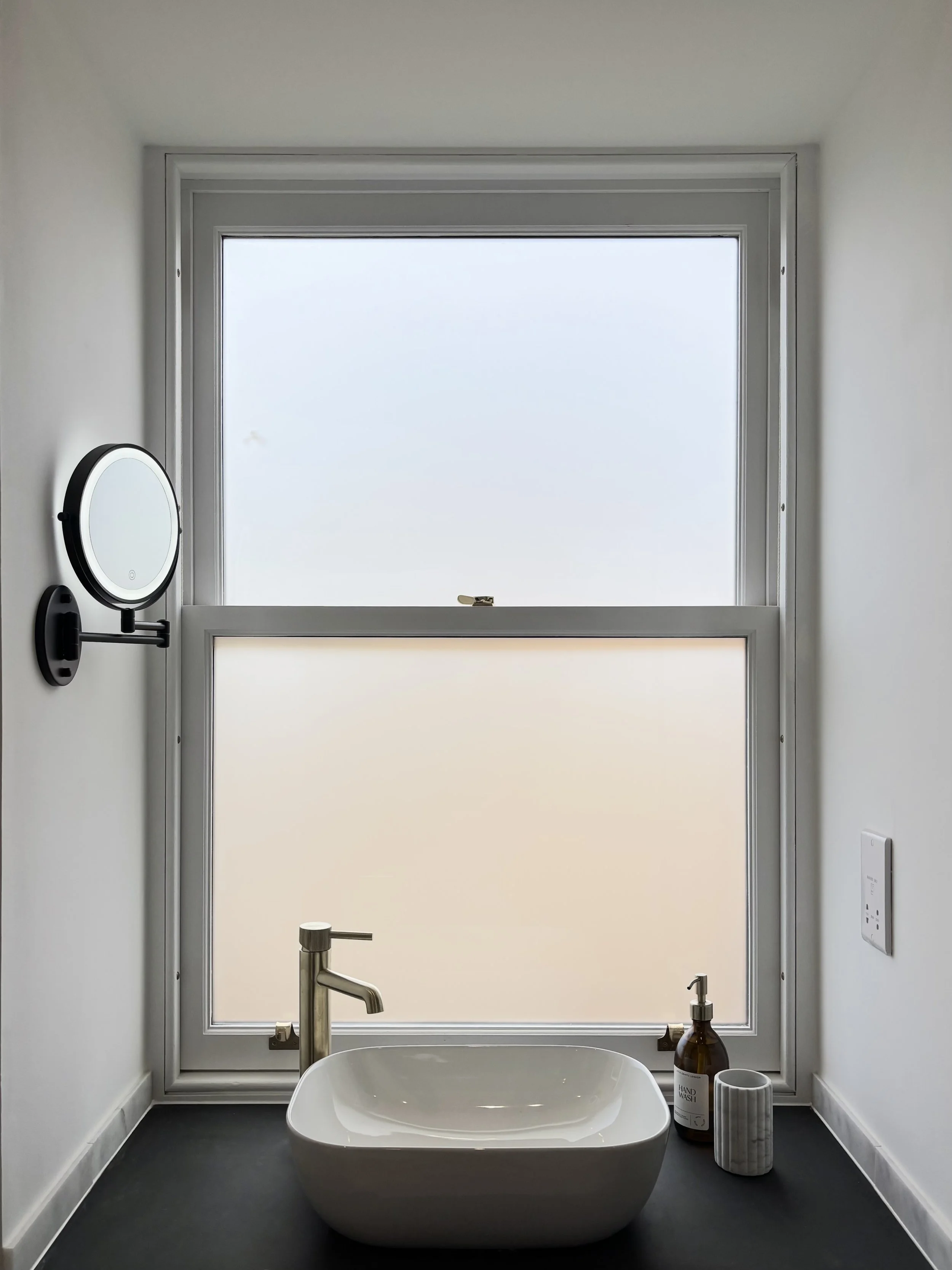 Modern bathroom with a white bathtub, large frosted window, and minimal decor including soap and hand wash bottles, and a mirror on the left wall.